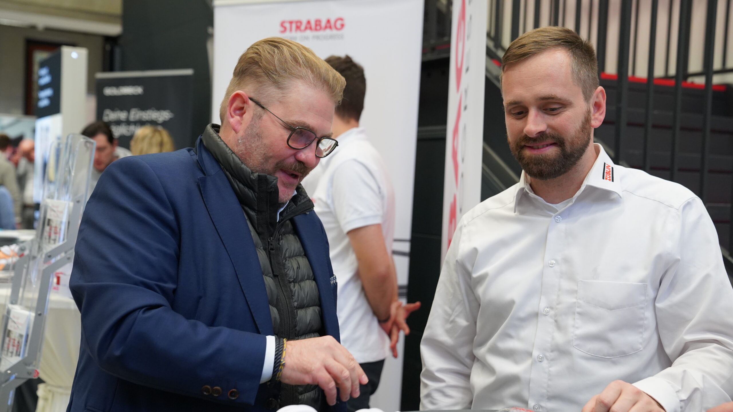 Zwei Männer unterhalten sich an einem Stand während einer Treff-Veranstaltung. Der eine trägt eine blaue Jacke und eine Brille, der andere ein weißes Hemd mit einem STRABAG-Logo. Sie scheinen sich über Unternehmen und Nachwuchstalente zu unterhalten, mit Broschüren im Hintergrund. at Hochschule Coburg