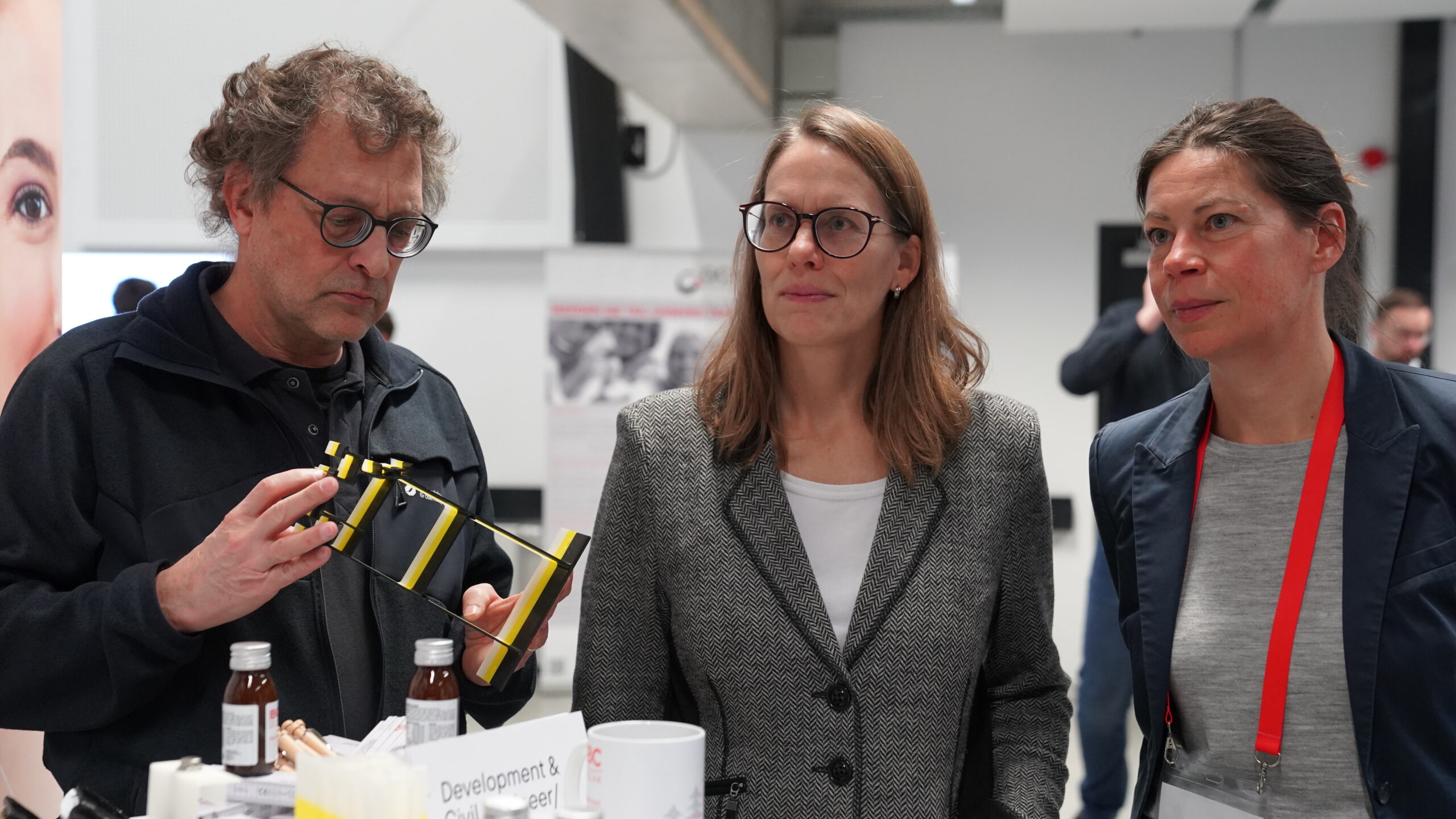 Drei Personen stehen in einem Innenraum eines Unternehmen-Treffs zusammen. Der Mann links hält einen gelb-schwarzen Gegenstand in der Hand, während die beiden Frauen neben ihm zugucken. Auf dem Tisch vor diesen Nachwuchstalenten sind Flaschen und Papiere ausgebreitet. at Hochschule Coburg