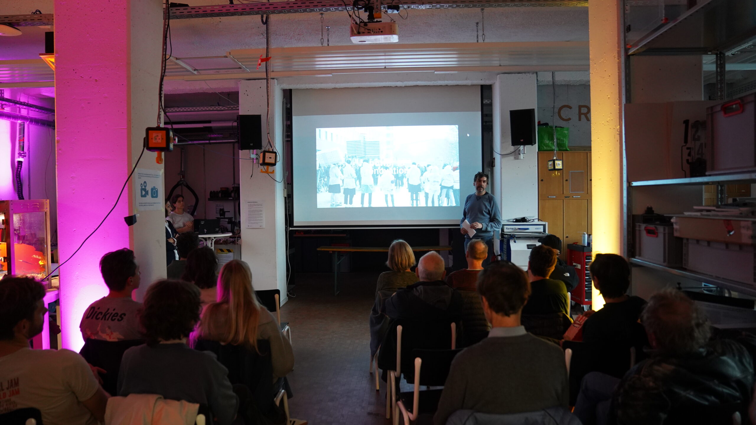 In einem schwach beleuchteten Raum der Hochschule sitzt eine Gruppe von Menschen vor einem Projektor und sieht sich eine Präsentation an. Der Vortragende steht vor einem Schwarz-Weiß-Bild, während rosa und gelbe Lichter die jüngste Renovierung des Raums hervorheben. at Hochschule Coburg