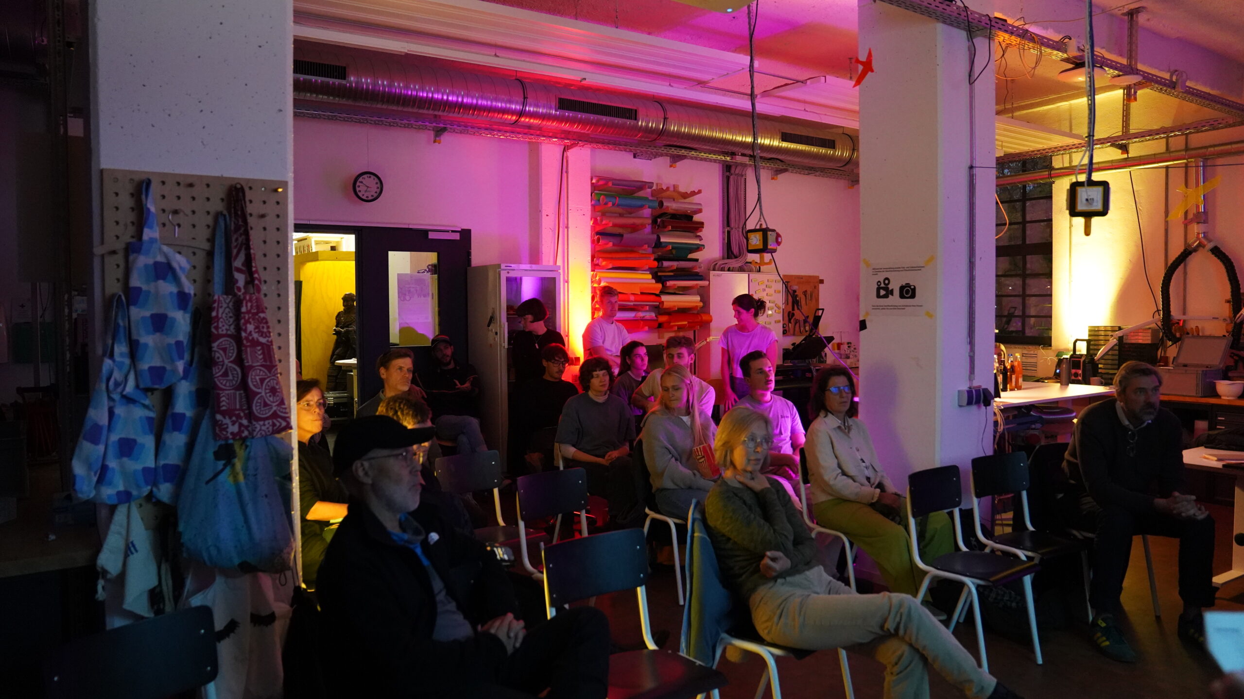 Eine Gruppe von Menschen sitzt auf Stühlen in einem schwach beleuchteten Raum und blickt nach vorne. Die bunte Beleuchtung, die Regale mit Gegenständen und die industrielle Ausstattung lassen auf eine Renovierung der Hochschule oder eine von Umbaukultur inspirierte Kreativwerkstatt schließen. at Hochschule Coburg