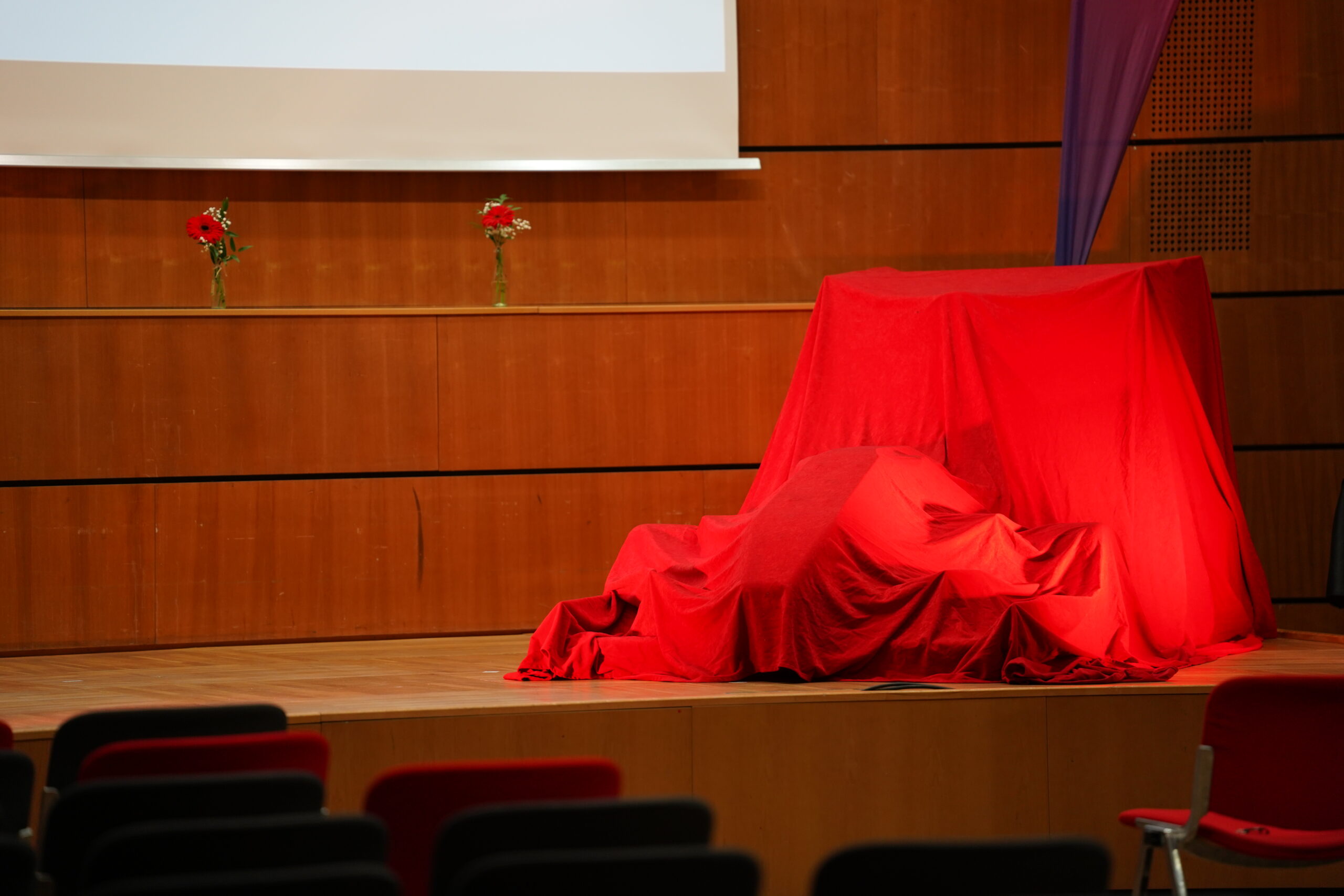 Auf einer Bühne mit einem hölzernen Hintergrund sind zwei mit leuchtend roten Tüchern bedeckte Objekte zu sehen, bei denen es sich möglicherweise um Leichtbau-Innovationen handelt, sowie drei rote Blumen in Vasen auf einem schmalen Regal darüber. Im Vordergrund sind einige leere Stühle zu sehen. at Hochschule Coburg