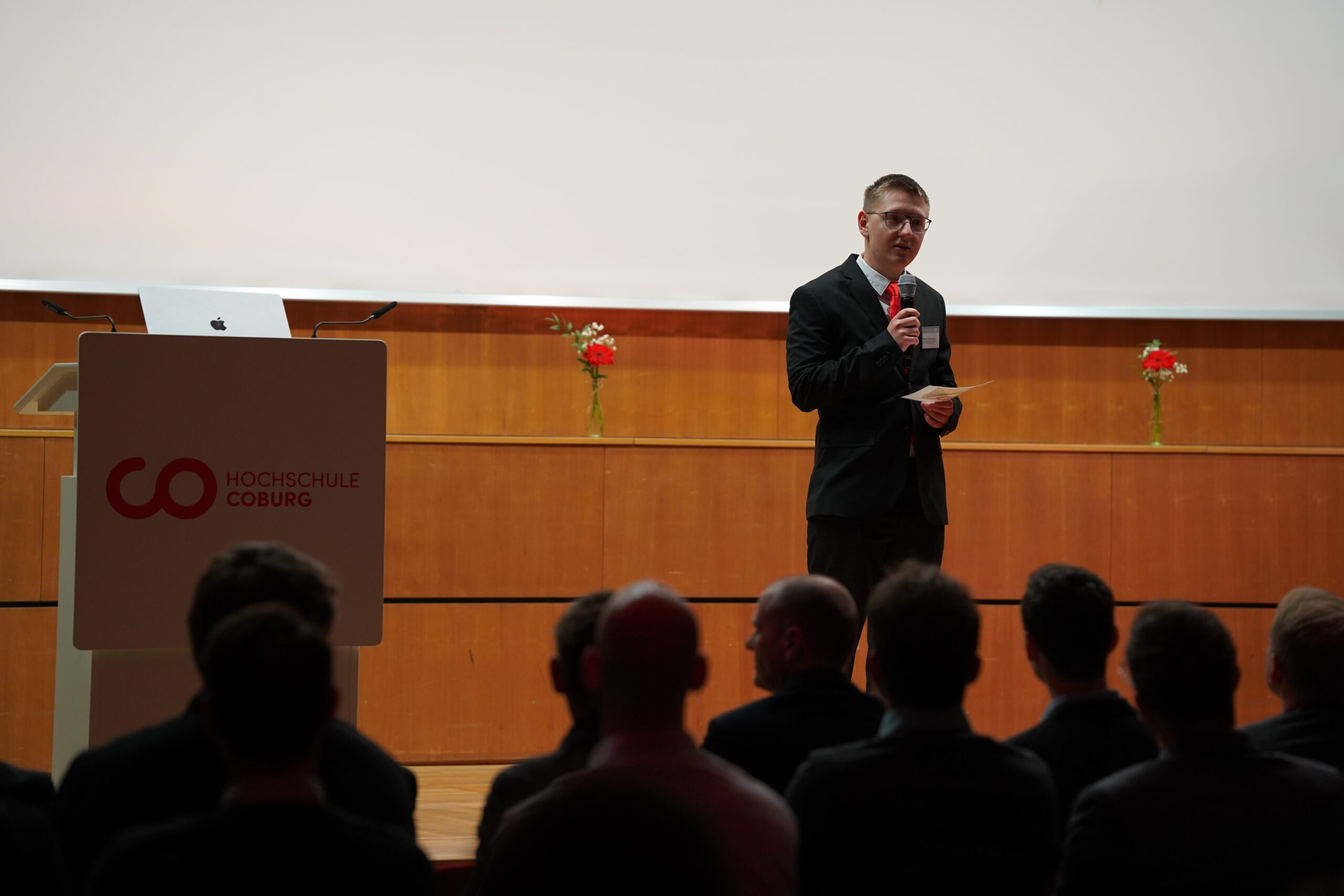 Ein Mann im Anzug spricht auf der Bühne über Leichtbau an der Hochschule Coburg, Mikrofon und Papier in der Hand. Silhouettierte Zuhörer schauen ihm zu. Im Hintergrund sind ein Podium, ein Laptop und zwei Vasen mit Blumen zu sehen. at Hochschule Coburg