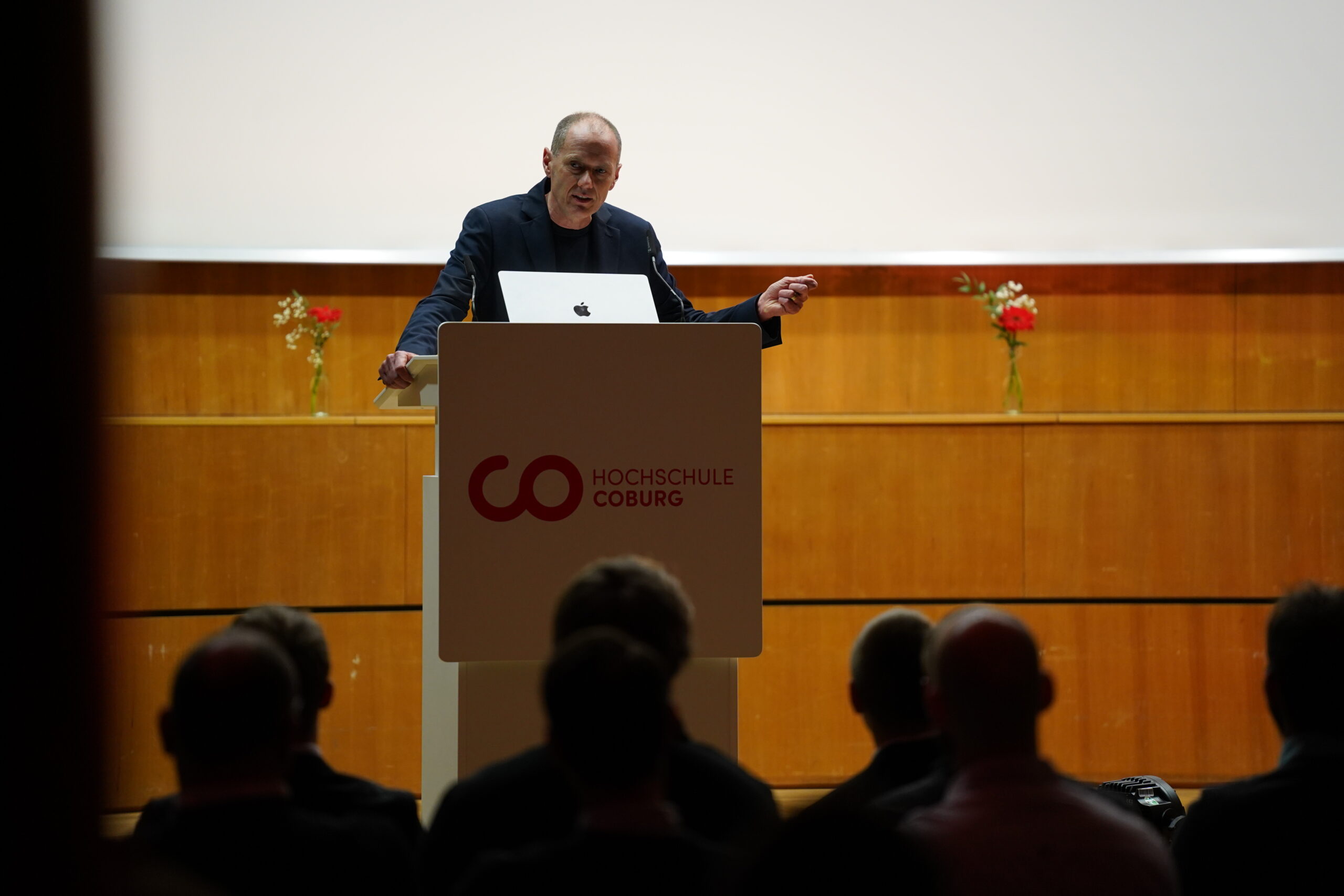 Eine Person steht an einem Podium und hält vor einem Publikum in einem Hörsaal der Hochschule Coburg einen Vortrag über Leichtbau, während im Hintergrund Blumen auf einem Holzsims angeordnet sind. at Hochschule Coburg