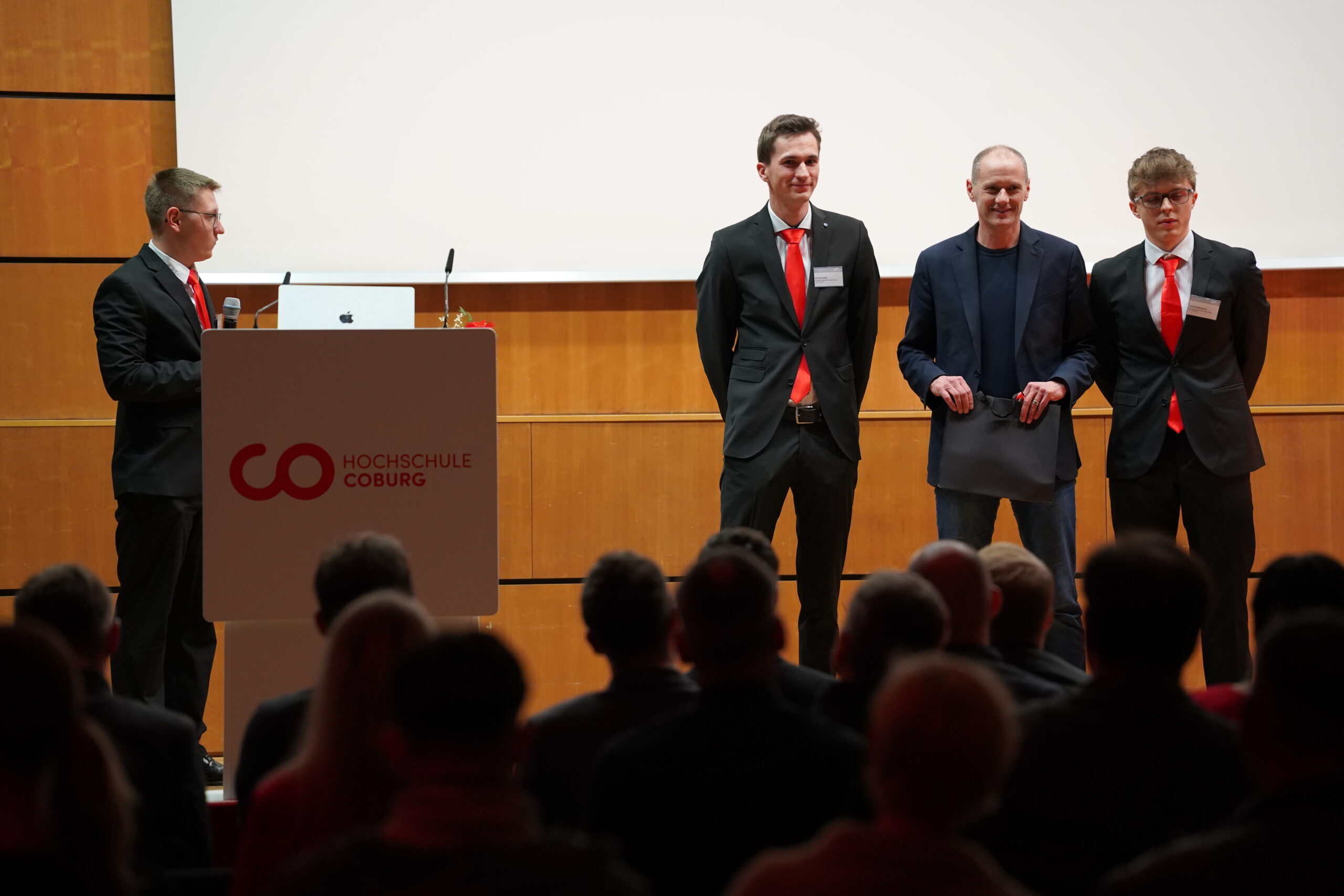 Vier Männer stehen bei einer Veranstaltung der Hochschule Coburg auf der Bühne und präsentieren Themen wie Aerodynamik und Leichtbau. Drei tragen schwarze Anzüge mit roten Krawatten, einer ein blaues Jackett. Ein Mann steht mit einem Laptop auf dem Podium und wendet sich an das aufmerksame Publikum. at Hochschule Coburg