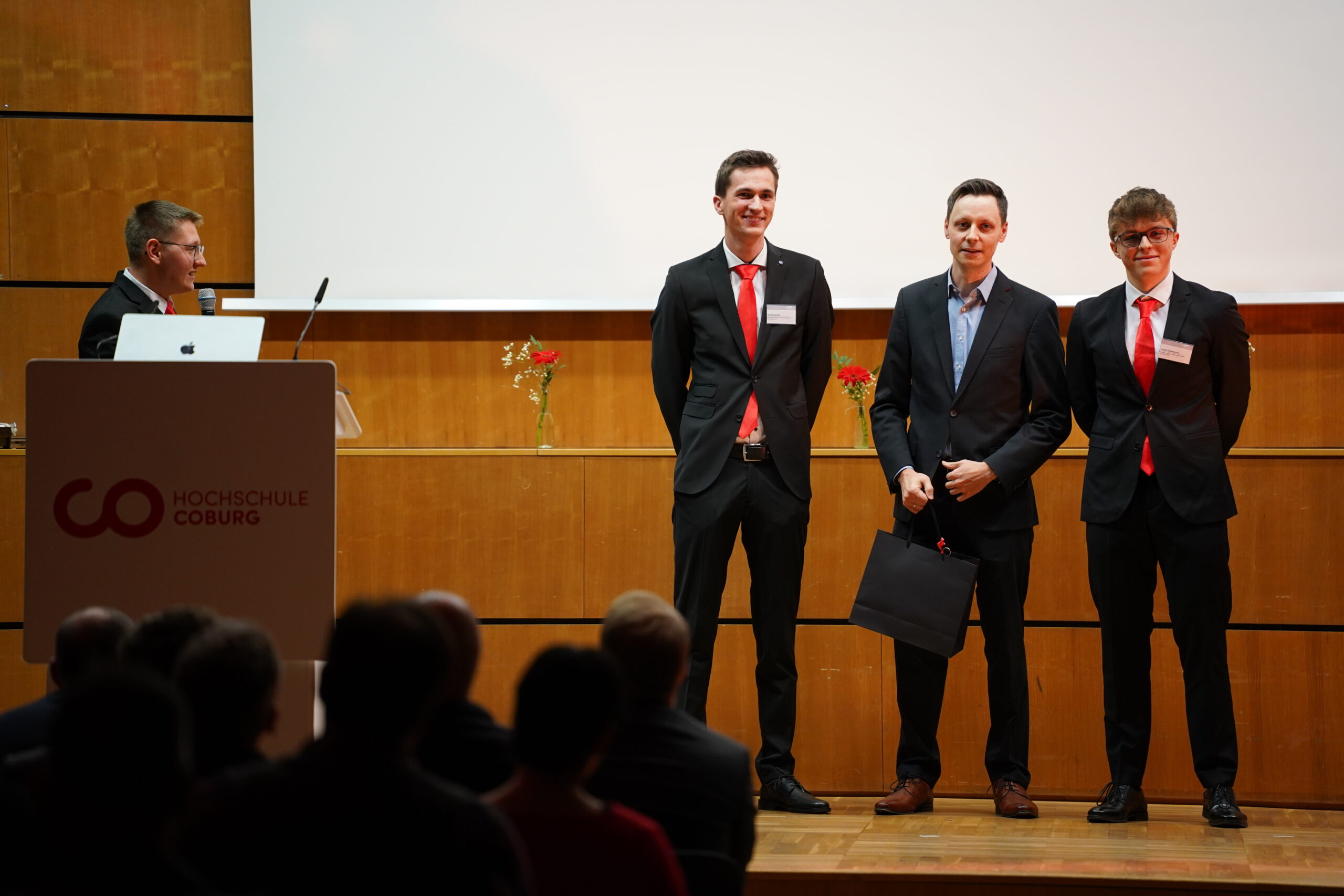 Vier Männer in Anzügen stehen auf einer Bühne der Hochschule Coburg. Drei wenden sich dem Publikum zu - zwei mit roten Krawatten, einer hält eine schwarze Geschenktüte in der Hand. Der vierte spricht an einem Podium mit einem Laptop über Themen wie Aerodynamik und Leichtbau. at Hochschule Coburg