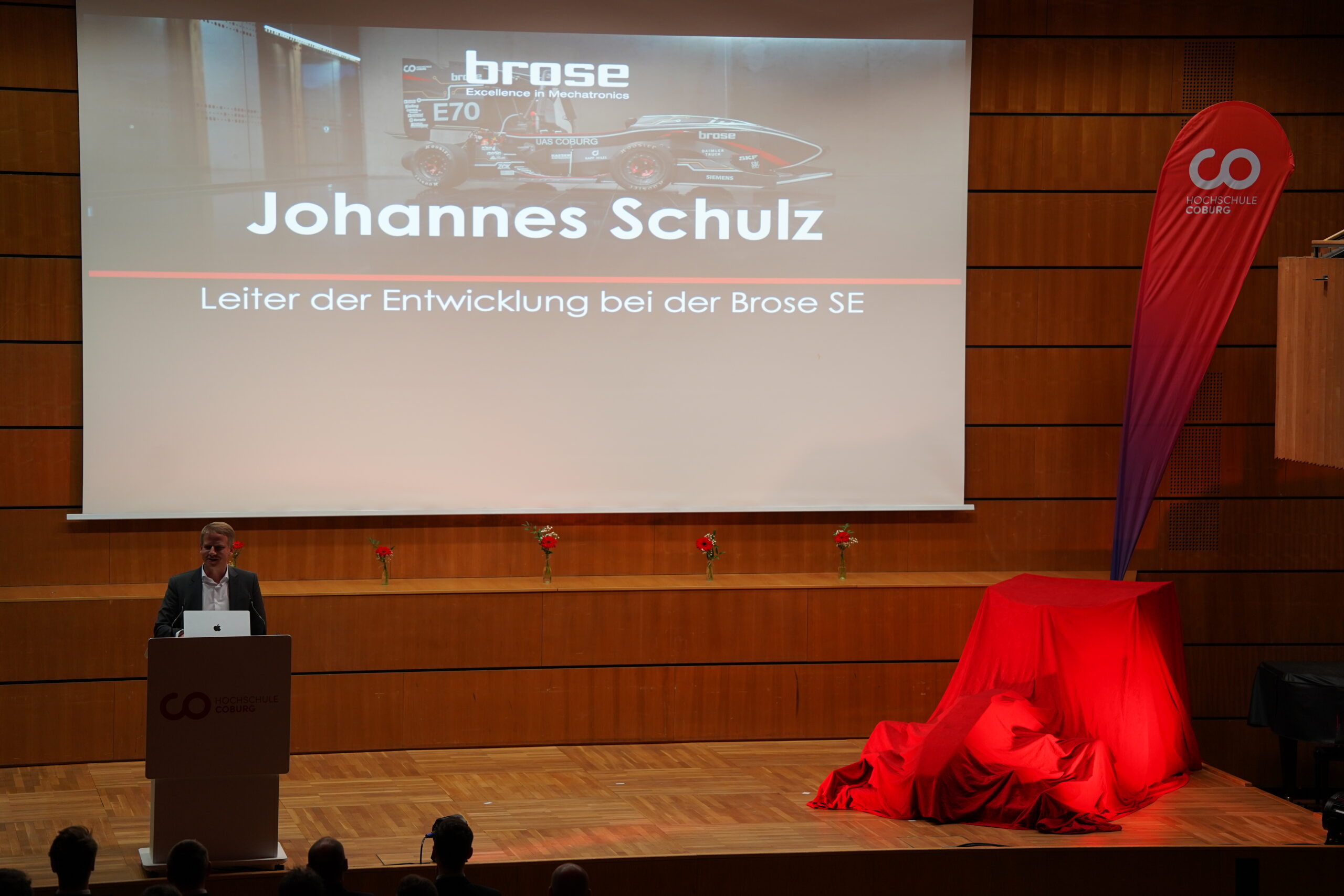 Ein Mann steht auf einem Podium und präsentiert in einem Hörsaal. Auf der Leinwand hinter ihm ist "Johannes Schulz, Leiter der Entwicklung bei der Brose SE" zu sehen - ein Bild eines Autos, das den neuesten Stand von Leichtbau und innovativer Elektrik verdeutlicht. Auf der Bühne stehen ein rot verhülltes Objekt und ein Transparent. at Hochschule Coburg