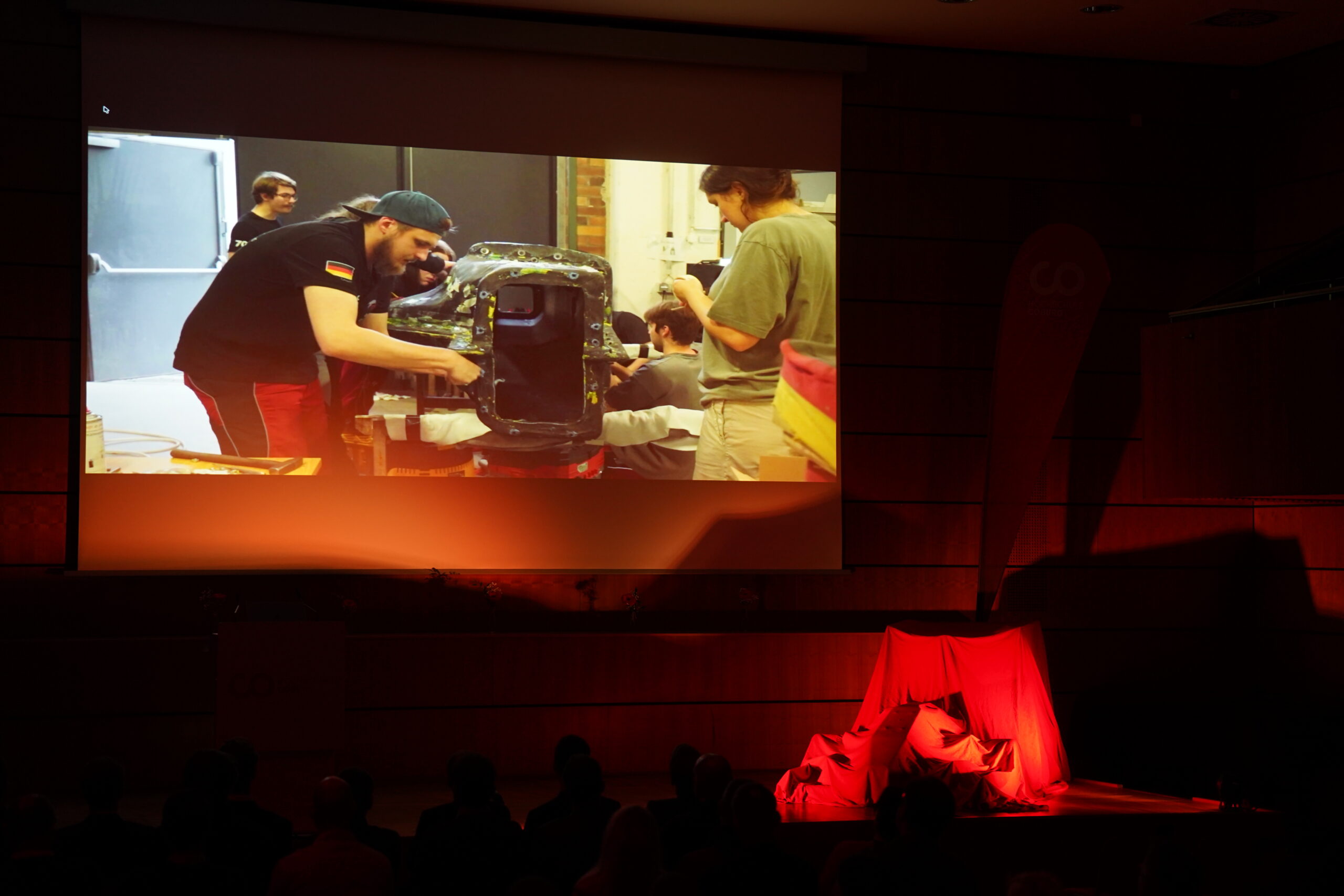 Eine Gruppe von Personen betrachtet die Projektion einer Werkstattszene zum Thema Leichtbau und Elektrik, die zwei Männer bei der Arbeit an einer Maschine zeigt, während auf der schwach beleuchteten Bühne unter der Leinwand eine mit rotem Stoff bedeckte Fläche zu sehen ist. at Hochschule Coburg