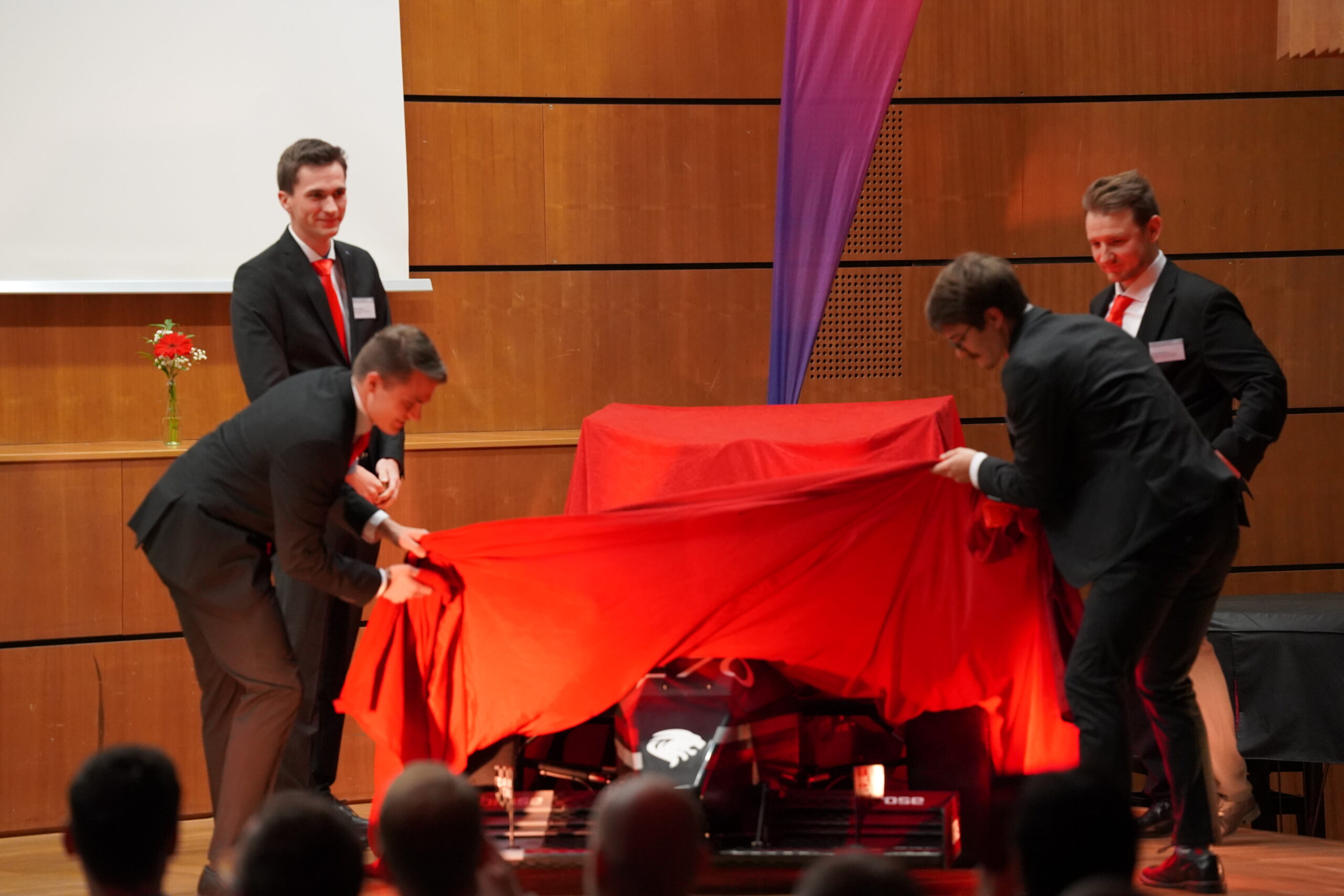 Vier Männer in Anzügen enthüllen auf der Bühne ein rotes Tuch, das ein Fahrzeug - möglicherweise einen Rennwagen - bedeckt und die Fortschritte in der Aerodynamik und im Leichtbau verdeutlicht. Im Hintergrund sind Holzverkleidungen und eine Blume in einer Vase auf einem Tisch zu sehen. at Hochschule Coburg