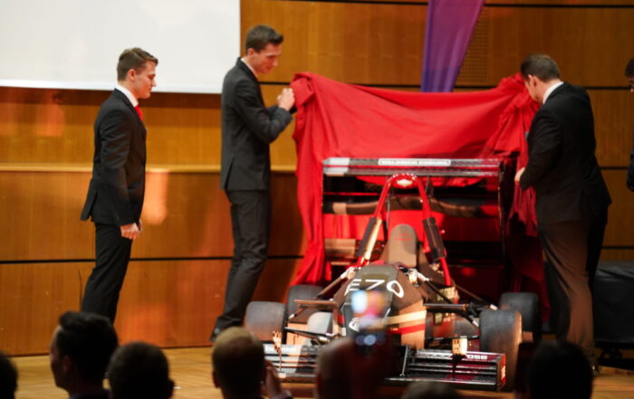 Drei Männer in Anzügen enthüllen auf einer Holzbühne vor Publikum einen schwarz-roten Formula Student-Rennwagen mit fortschrittlicher Aerodynamik und der Nummer E70 auf der Nase. at Hochschule Coburg