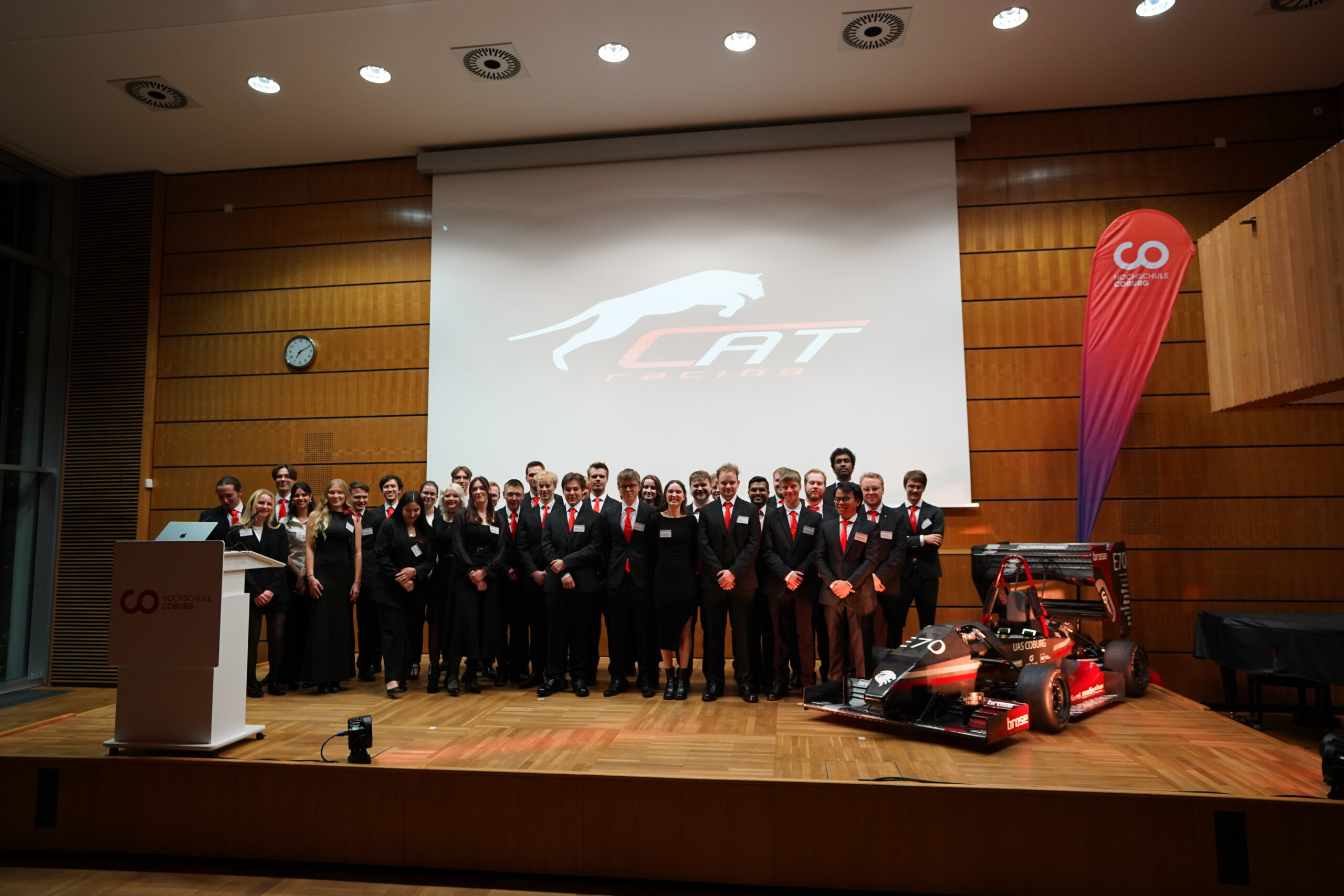 Eine Gruppe von formell gekleideten Personen steht auf einer Bühne vor einem Bildschirm, auf dem das Logo einer springenden Katze und der Schriftzug "CAT" zu sehen sind. Ein kleiner, aerodynamischer Rennwagen ist vor ihnen in dem modernen Zuschauerraum geparkt. at Hochschule Coburg
