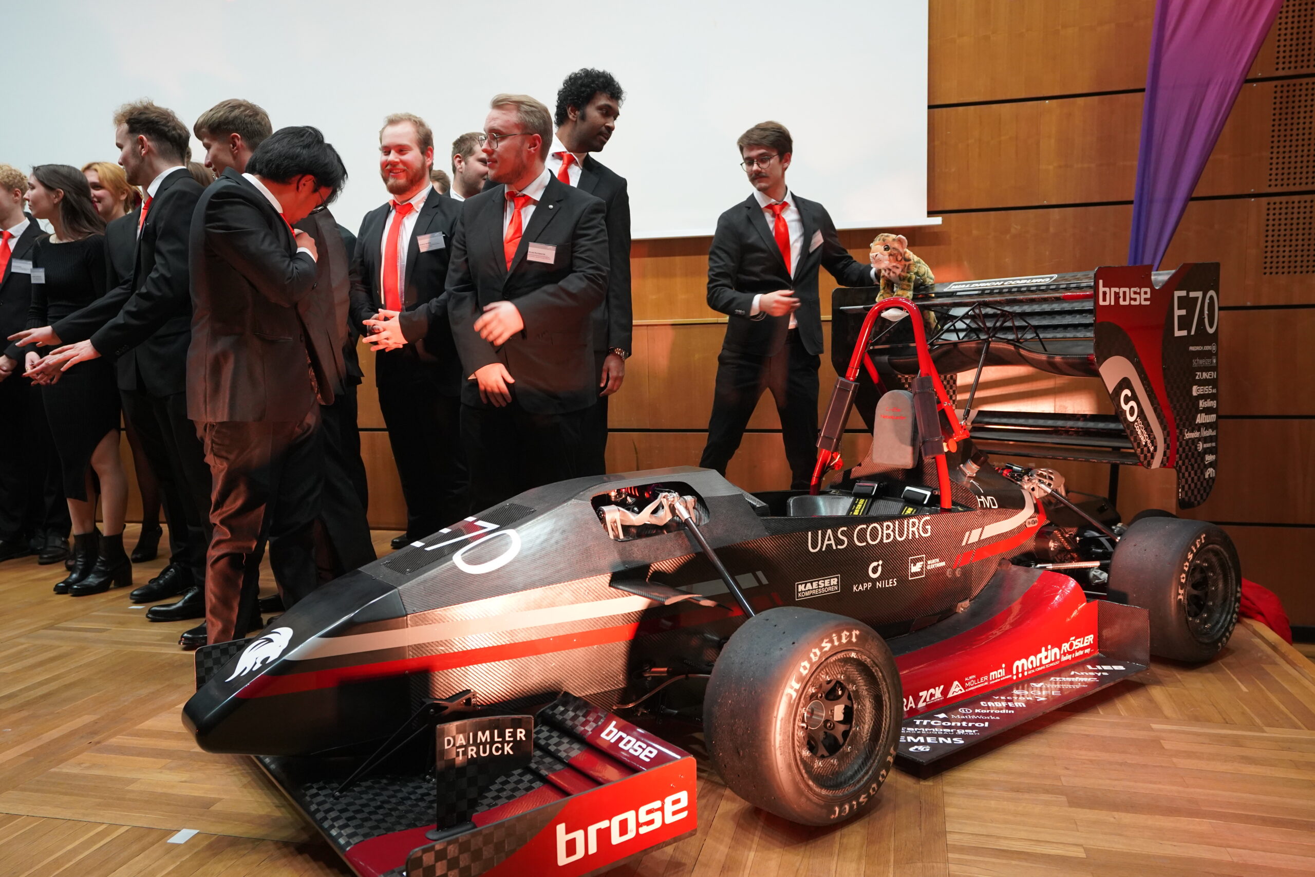 Eine Gruppe von Menschen in Anzügen und roten Krawatten steht auf einer hölzernen Bühne neben einem schwarz-roten Rennwagen mit der Aufschrift "FH Coburg" und "brose" und scheint die Leistungen des Teams in Leichtbau und Aerodynamik bei einer Indoor-Veranstaltung zu feiern. at Hochschule Coburg