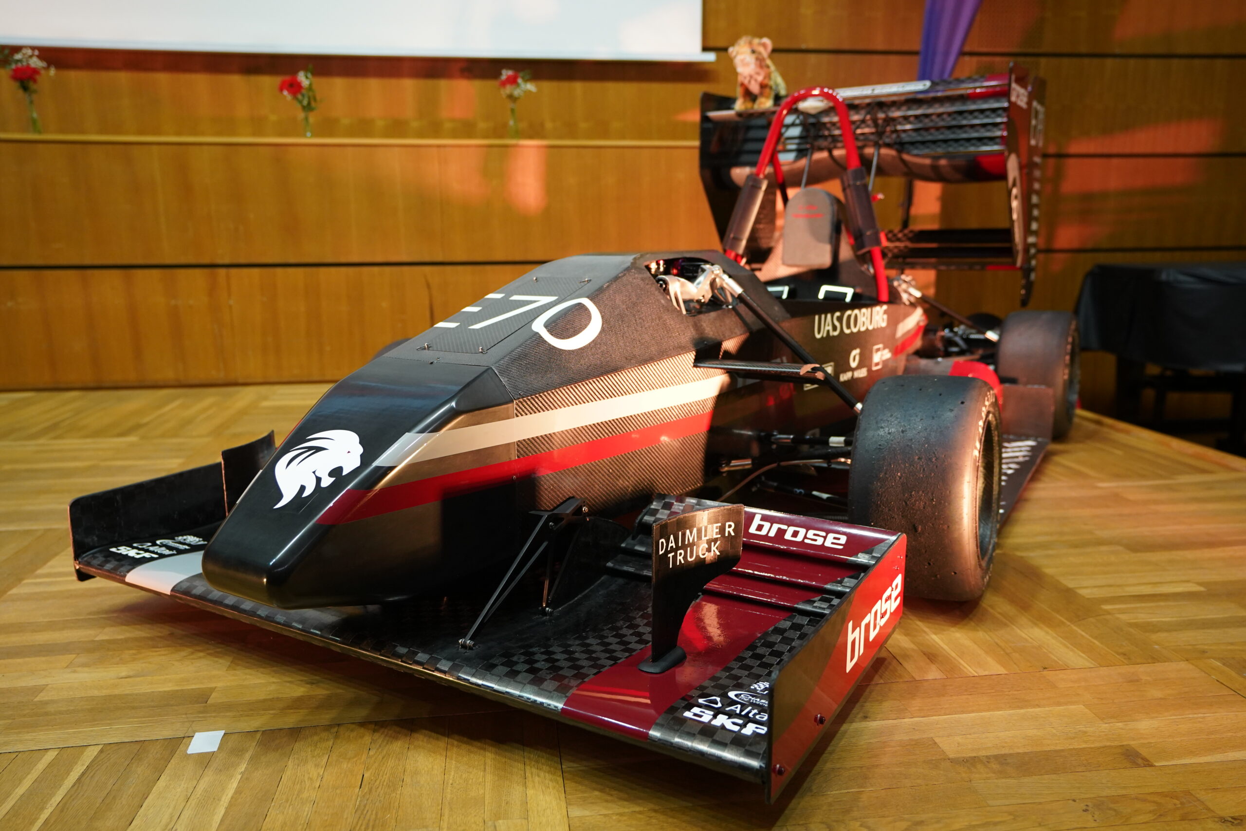 Ein schwarz-roter Formel-Rennwagen mit einem Löwen-Logo auf der Nase präsentiert seine Aerodynamik und seinen Leichtbau im Innenraum auf einem Holzboden vor einer holzgetäfelten Wand mit Blumen im Hintergrund. at Hochschule Coburg