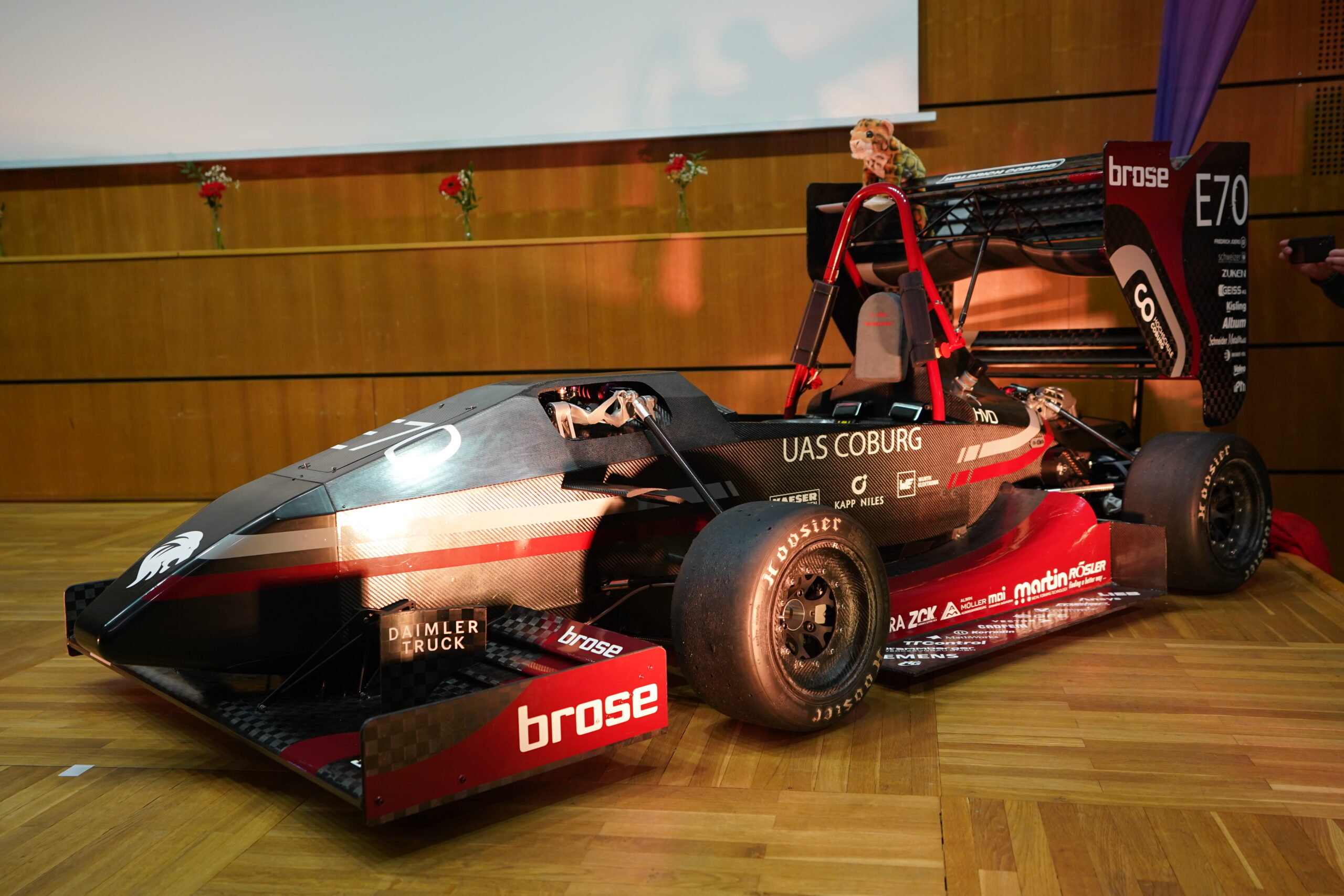 Ein schwarz-roter Formula Student-Rennwagen mit "UAS COBURG"- und Sponsorenlogos in fortschrittlichem Leichtbau-Design steht in einem Innenraum auf einem Holzboden. Auf dem Fahrersitz nimmt eine Puppe Platz. at Hochschule Coburg