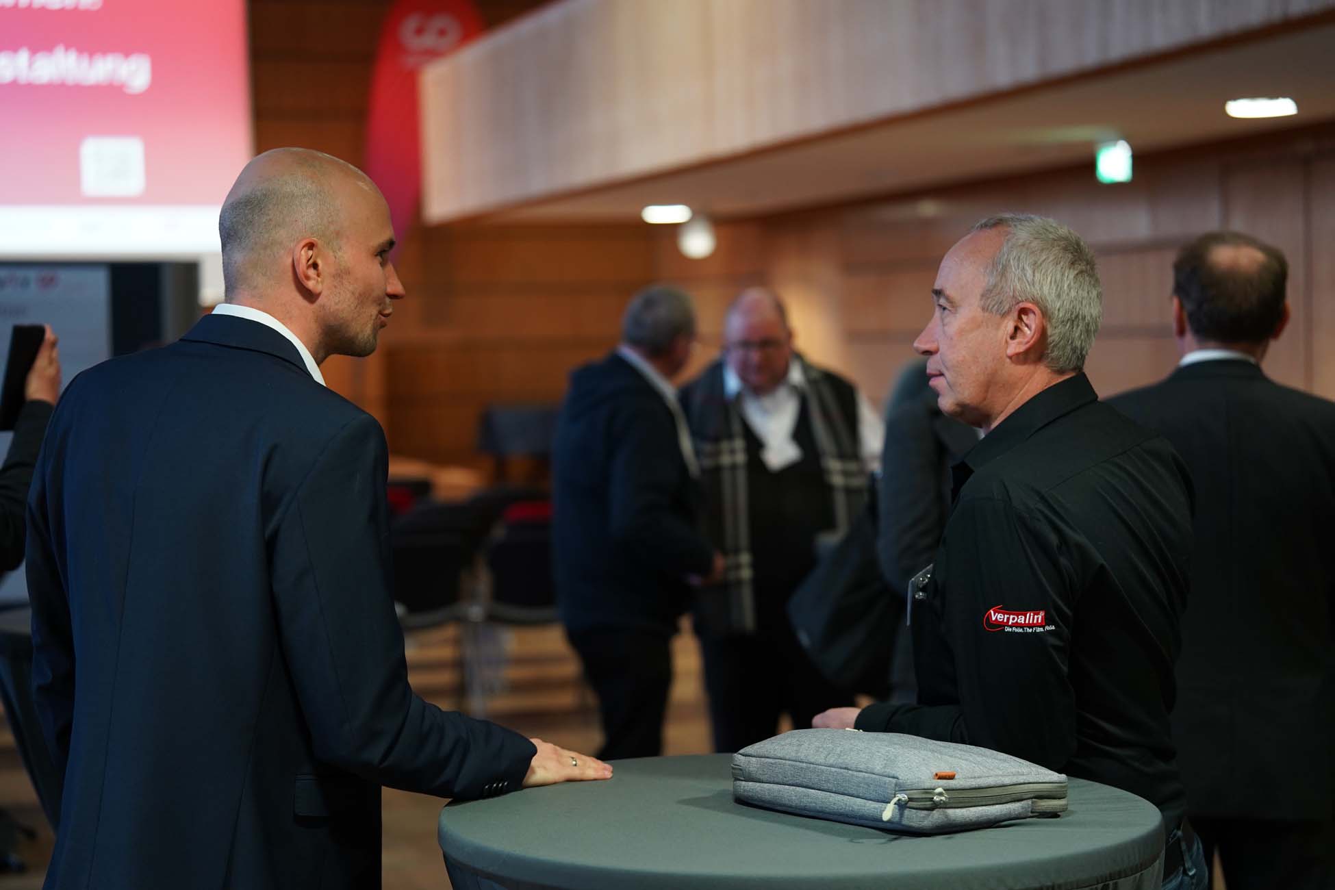 Zwei Männer unterhalten sich an einem runden Tisch in einem Konferenzsaal. Der eine trägt einen Anzug, der andere ein schwarzes Hemd mit dem Logo des Automatisch gespeicherten Entwurfs, während sich im Hintergrund mehrere Personen unterhalten. at Hochschule Coburg