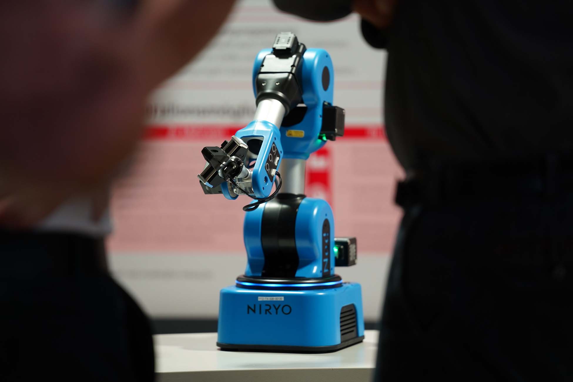 Ein blauer Roboterarm mit der Aufschrift "Niryo" steht auf einem Tisch, mit verschwommenen Menschen im Vordergrund und einem Plakat mit rotem Text im Hintergrund - ein automatisch gespeicherter Entwurf für fortschrittliche Robotik. at Hochschule Coburg