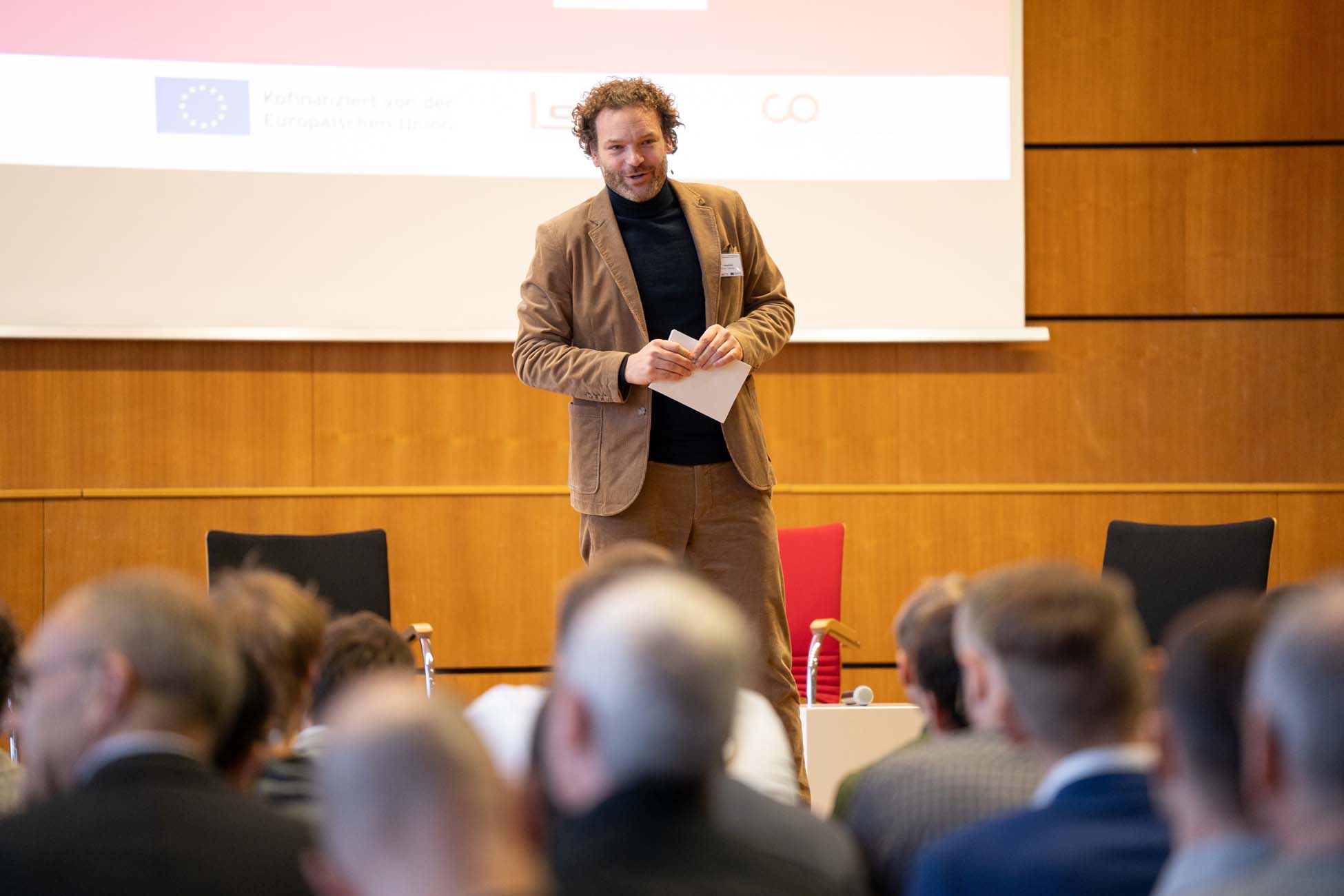 Ein Mann in einem braunen Anzug steht auf der Bühne vor einem Publikum und hält Papiere in der Hand. Hinter ihm zeigt eine Projektionsfläche Logos und Texte, darunter "Automatisch gespeicherter Entwurf". Der Raum ist holzgetäfelt und auf der Bühne sind Stühle zu sehen. at Hochschule Coburg