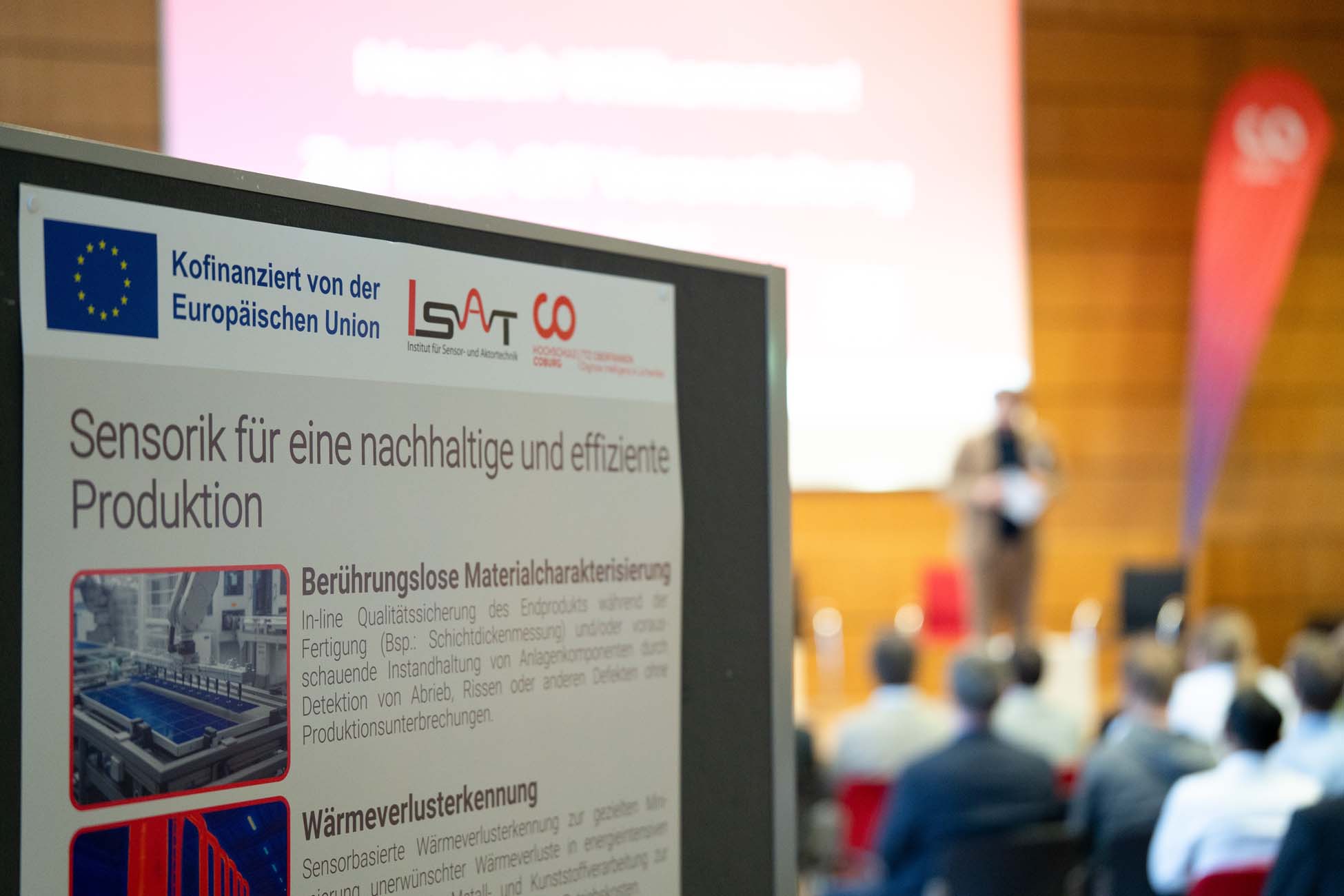 Im Mittelpunkt steht ein Plakat über nachhaltige und effiziente Produktion, das als automatisch gespeicherter Entwurf gekennzeichnet und mit dem EU-Logo versehen ist. Im unscharfen Hintergrund spricht eine Person auf der Bühne zu einem Publikum in einer Konferenzumgebung. at Hochschule Coburg