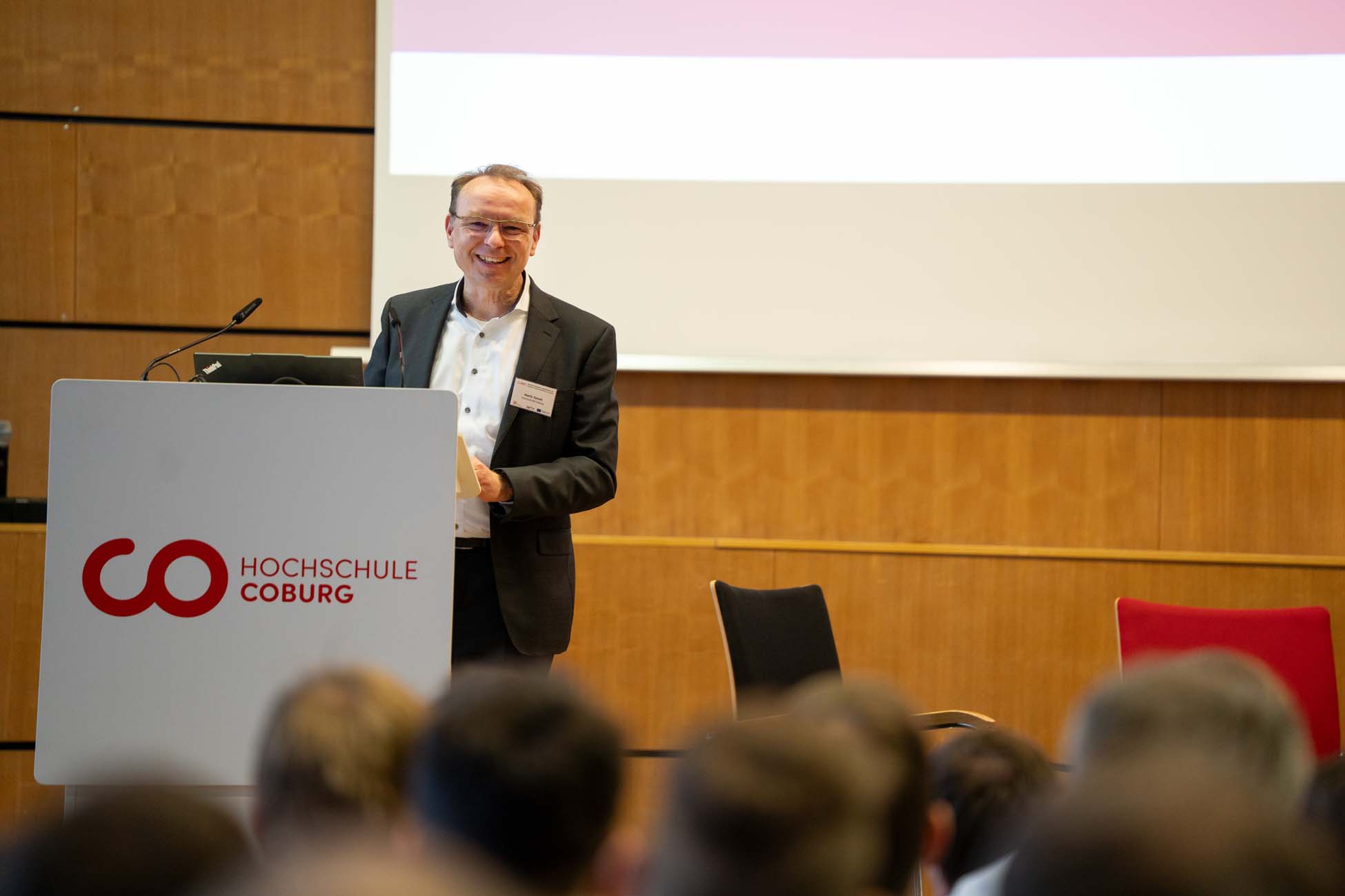 Ein Mann im Anzug steht an einem Podium und hält einen Vortrag vor einem Publikum an der Hochschule Coburg. Hinter ihm ist ein großer Bildschirm zu sehen, auf dem ein automatisch gespeicherter Entwurf angezeigt wird, und im Vordergrund sitzen Menschen. at Hochschule Coburg