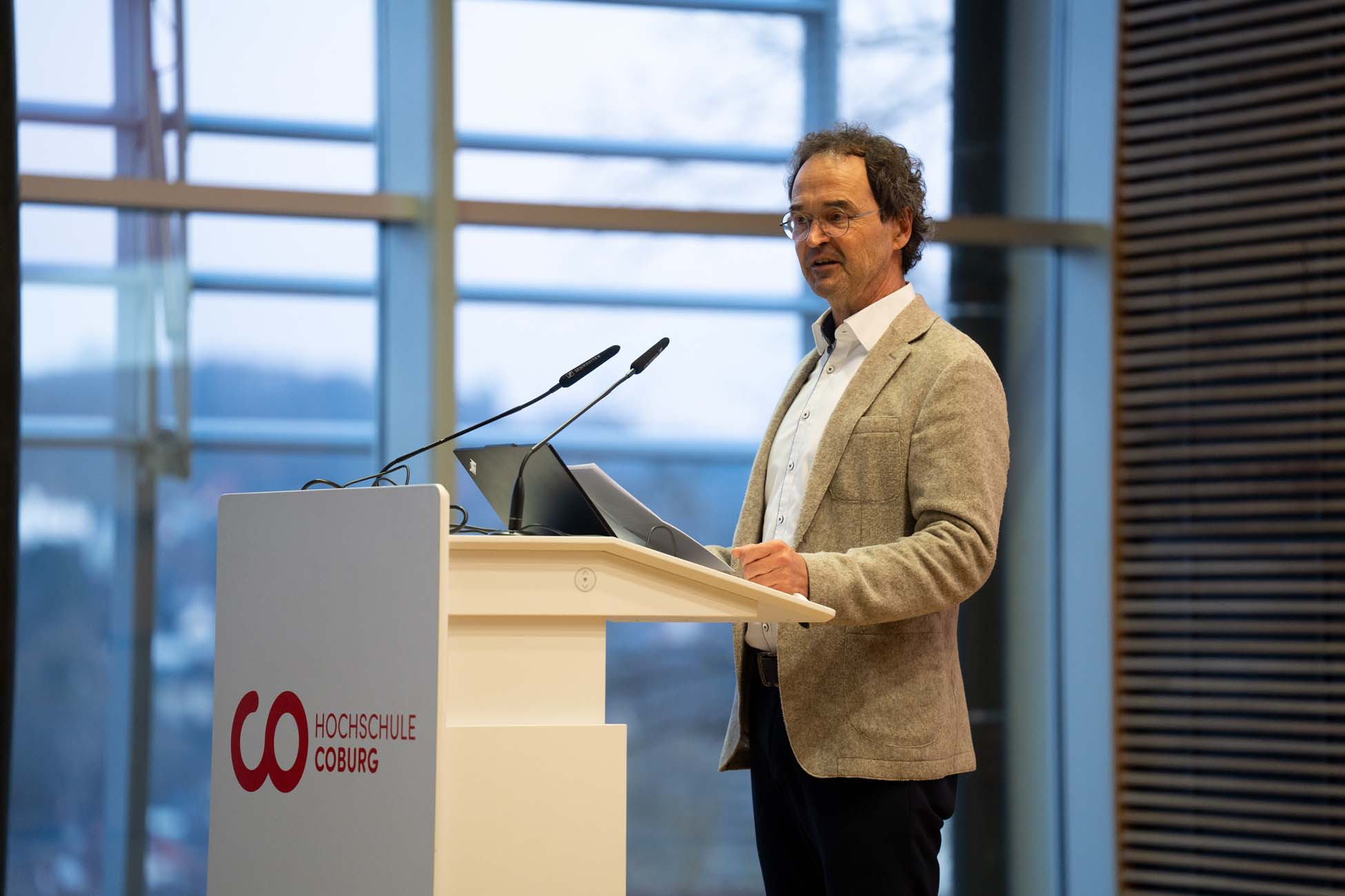 Ein Mann in beiger Jacke und weißem Hemd steht an einem Podium mit Mikrofonen und einem Laptop und hält einen automatisch gespeicherten Entwurf an der Hochschule Coburg. Im Hintergrund sind große Fenster mit vertikalen Jalousien zu sehen. at Hochschule Coburg