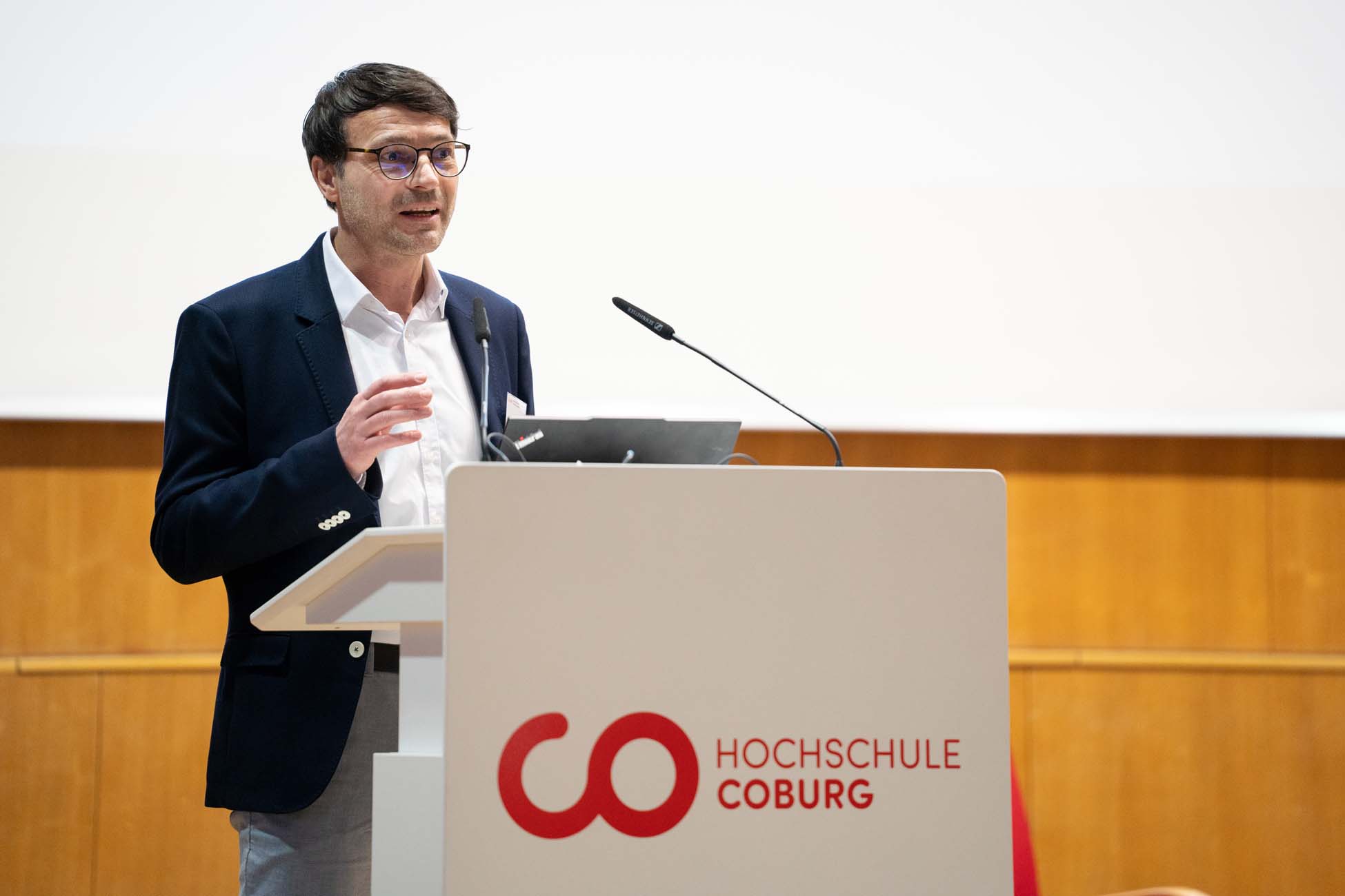 Ein Mann in einer Anzugsjacke spricht an einem Podium mit einem Mikrofon, neben einem automatisch gespeicherten Entwurf. Das Podium zeigt das Logo der Hochschule Coburg vor einer Holzwand und einem weißen Bildschirm. at Hochschule Coburg
