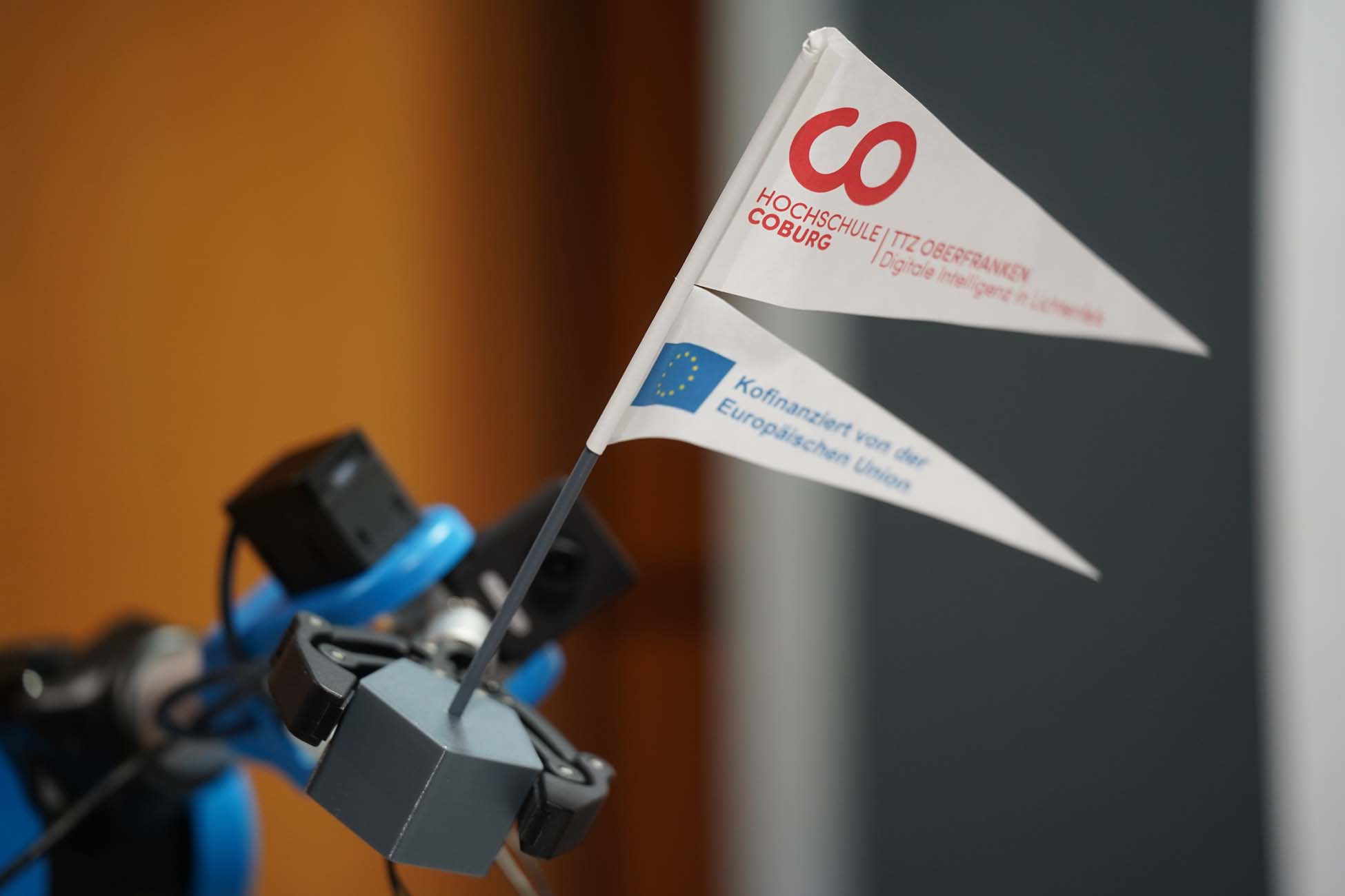Eine Nahaufnahme von zwei kleinen Fahnen an einem Roboterarm, die automatisch als Entwurf erfasst wurden, eine mit dem Logo der Hochschule Coburg und die andere mit dem Hinweis auf die EU-Kofinanzierung, vor einem unscharfen Hintergrund. at Hochschule Coburg