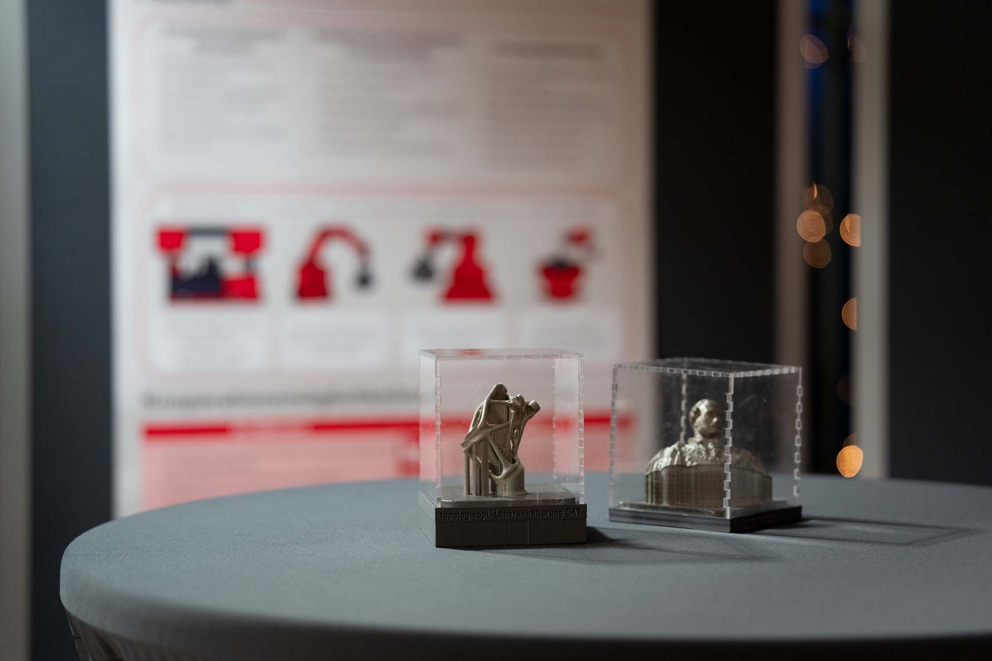 Zwei kleine 3D-gedruckte Figuren stehen in durchsichtigen Plastikboxen auf einem runden grauen Tisch, im Hintergrund ein verschwommenes Informationsplakat und ein automatisch gespeicherter Entwurf. at Hochschule Coburg