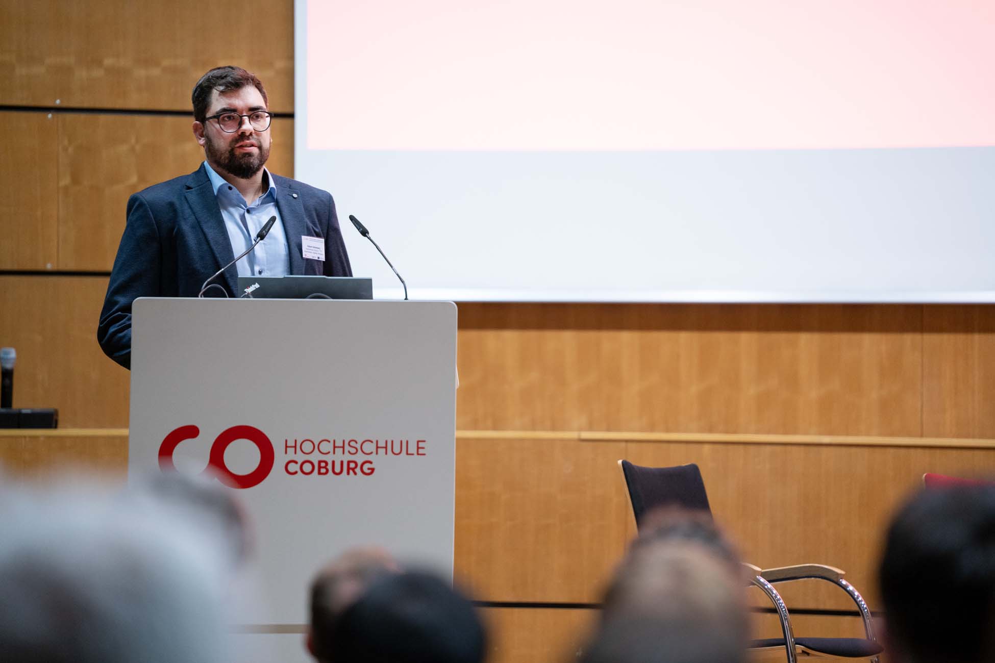 Ein Mann im Anzug steht an einem Podium mit der Aufschrift "Hochschule Coburg" und spricht in einem Hörsaal in Mikrofone. Die Szene, die einem automatisch gespeicherten Entwurf ähnelt, umfasst eine Projektionsfläche und einen leeren Stuhl im Hintergrund. at Hochschule Coburg