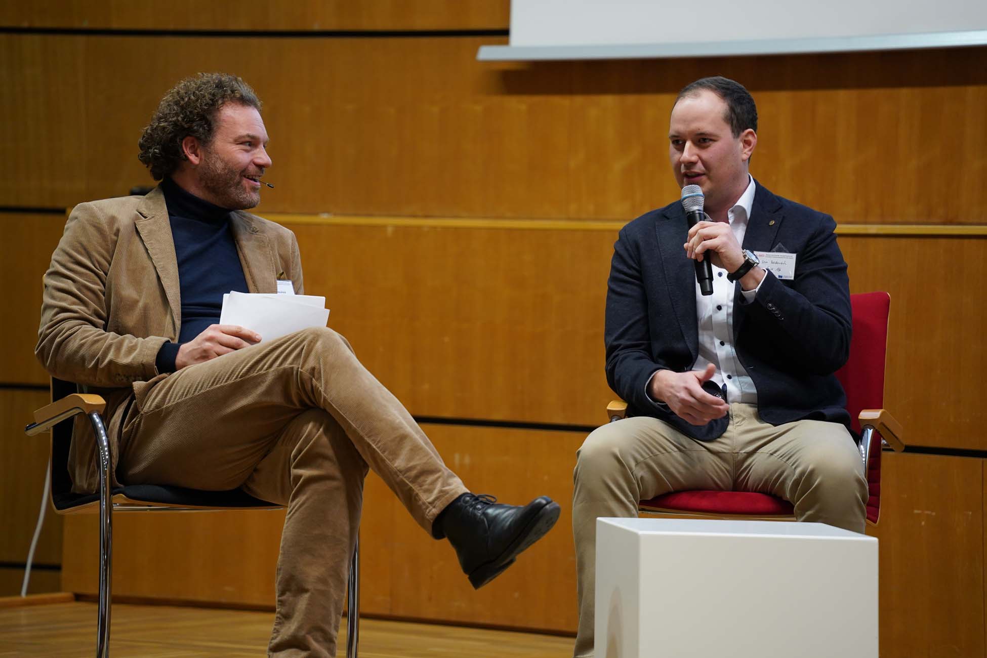 Zwei Männer sitzen auf der Bühne und führen eine Diskussion. Der eine hält ein Klemmbrett und lächelt zu einem automatisch gespeicherten Entwurf, während der andere ein Mikrofon hält und spricht. Beide tragen Blazer und sitzen vor einer Holzwand. at Hochschule Coburg