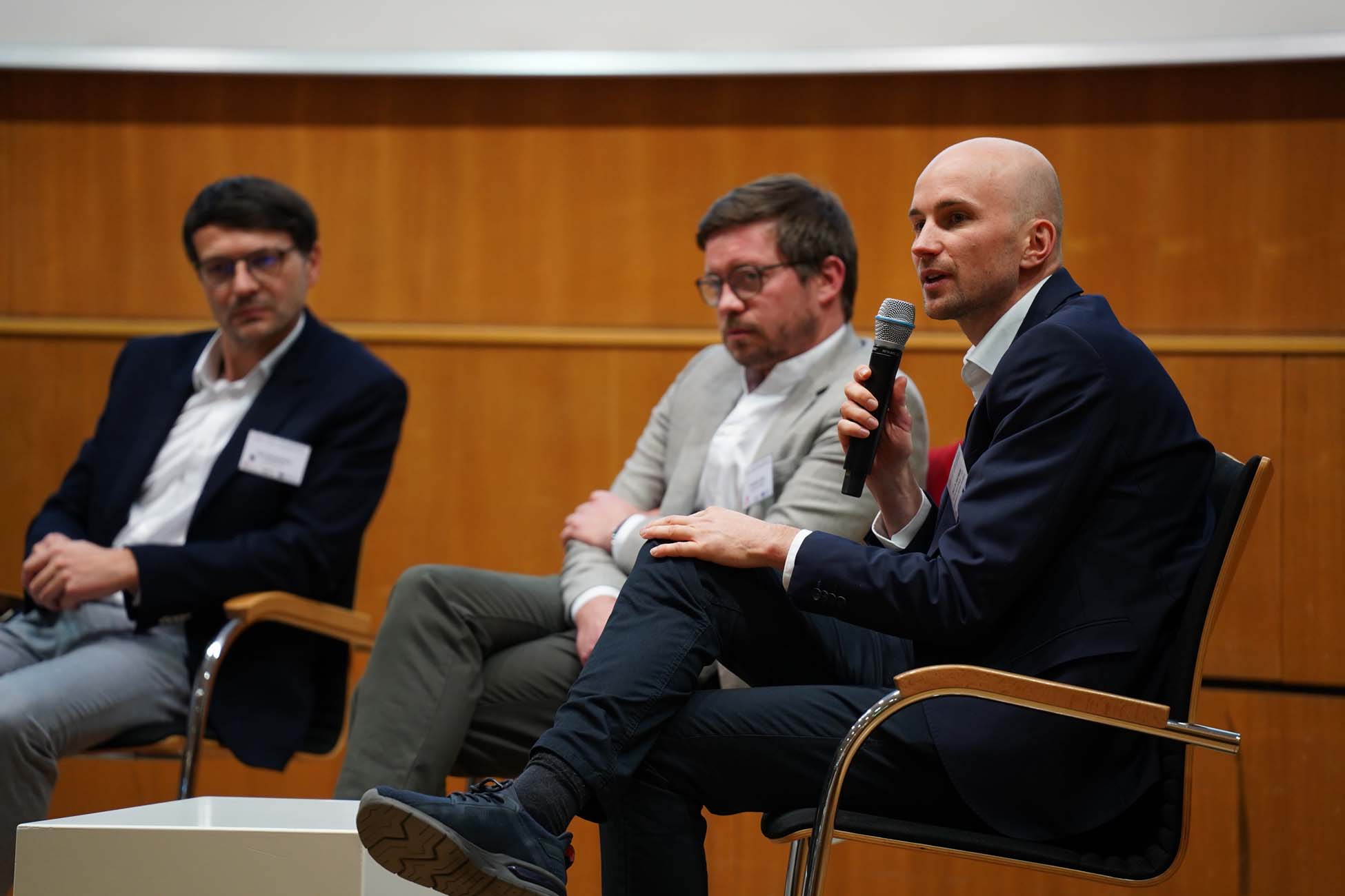 Drei Männer in Geschäftskleidung sitzen auf Stühlen in einer Podiumsdiskussion, wobei ein Mann ein Mikrofon hält und spricht. Im Hintergrund sind eine Holzwand und eine Projektionsfläche mit der Aufschrift "Automatisch gespeicherter Entwurf" zu sehen. at Hochschule Coburg