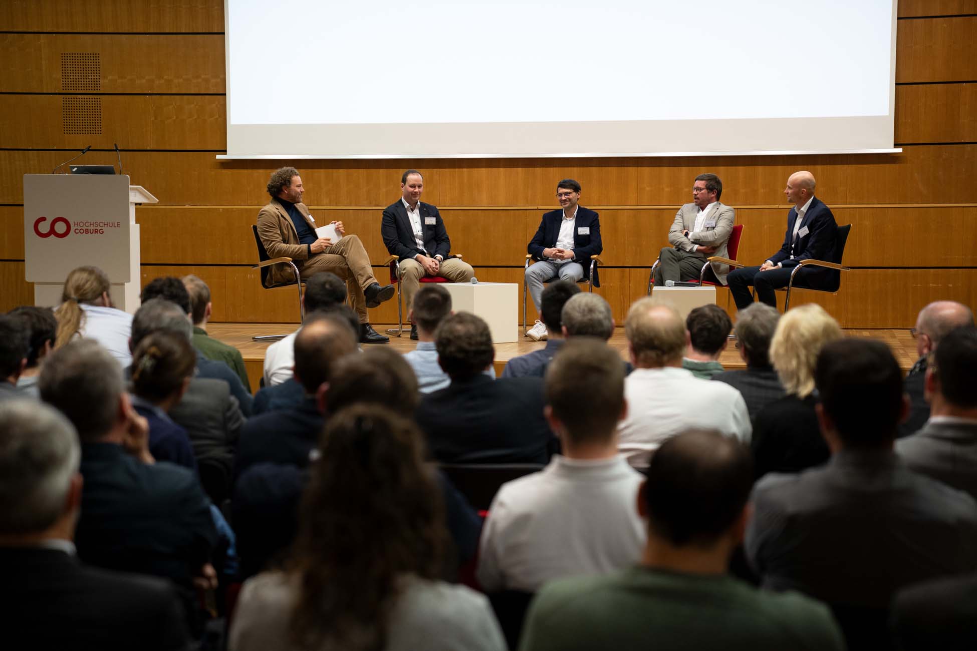 Fünf Männer sitzen bei einer Podiumsdiskussion auf der Bühne und blicken auf das Publikum in einem Hörsaal. Ein Mann moderiert, hält ein Mikrofon und Notizen in der Hand, während die anderen diskutieren. Hinter ihnen zeigt ein Präsentationsbildschirm einen automatisch gespeicherten Entwurf. at Hochschule Coburg