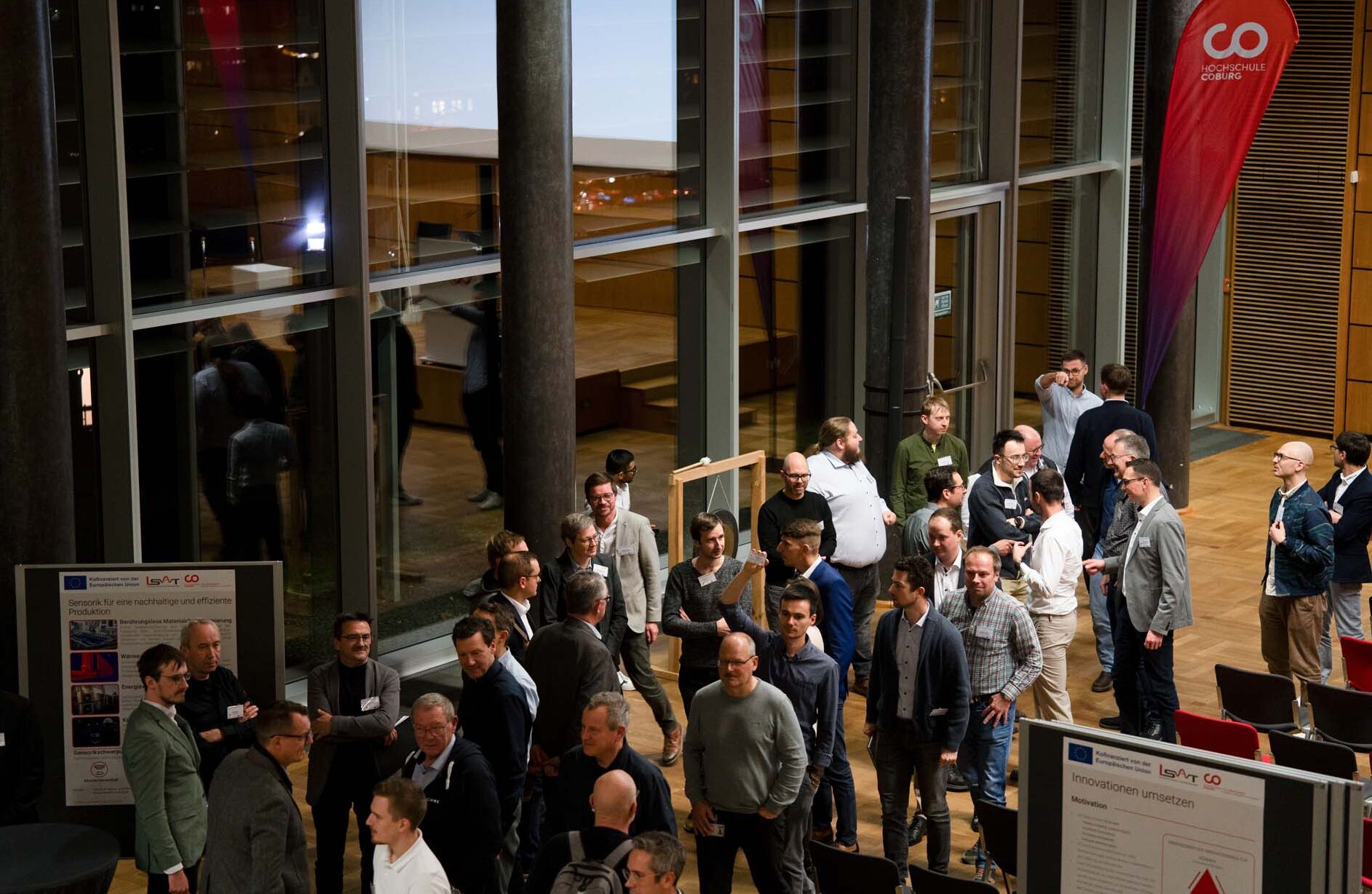Eine Gruppe von Menschen knüpft in einer geräumigen, modernen Halle mit großen Fenstern Kontakte und Kontakte. Einige betrachten Informationsplakate, während andere in kleinen Gruppen in der Nähe eines automatisch gespeicherten Entwurfs und eines roten Veranstaltungsbanners stehen und sich unterhalten. at Hochschule Coburg
