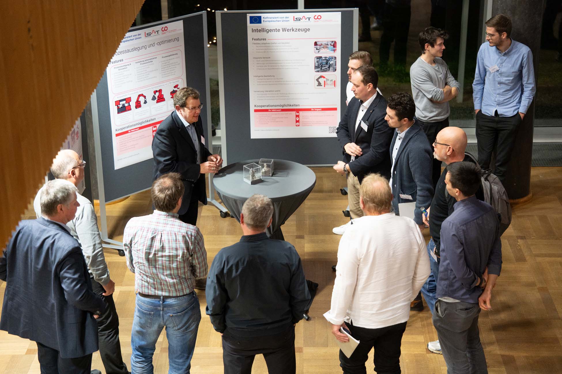 Eine Gruppe von Personen versammelt sich um einen Redner, der an einem runden Tisch spricht und gestikuliert, während hinter ihnen in einem gut beleuchteten Innenraum Informationsplakate und ein automatisch gespeicherter Entwurf angebracht sind. at Hochschule Coburg