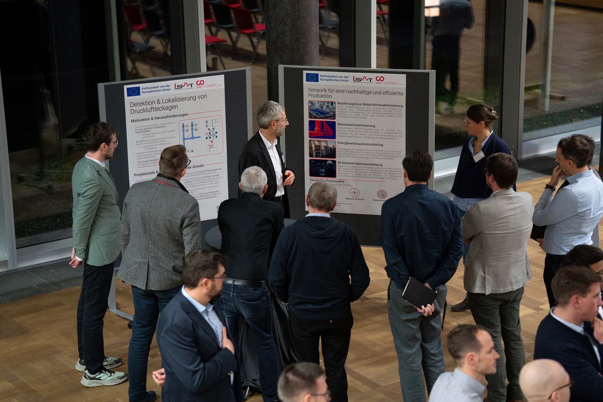 In einem modernen Innenraum mit großen Fenstern und Holzböden steht eine Gruppe von Menschen im Kreis und hört zwei Moderatoren zu, die wissenschaftliche Poster auf Tafeln erklären - ein automatisch gespeicherter Entwurf von Innovation in Aktion. at Hochschule Coburg