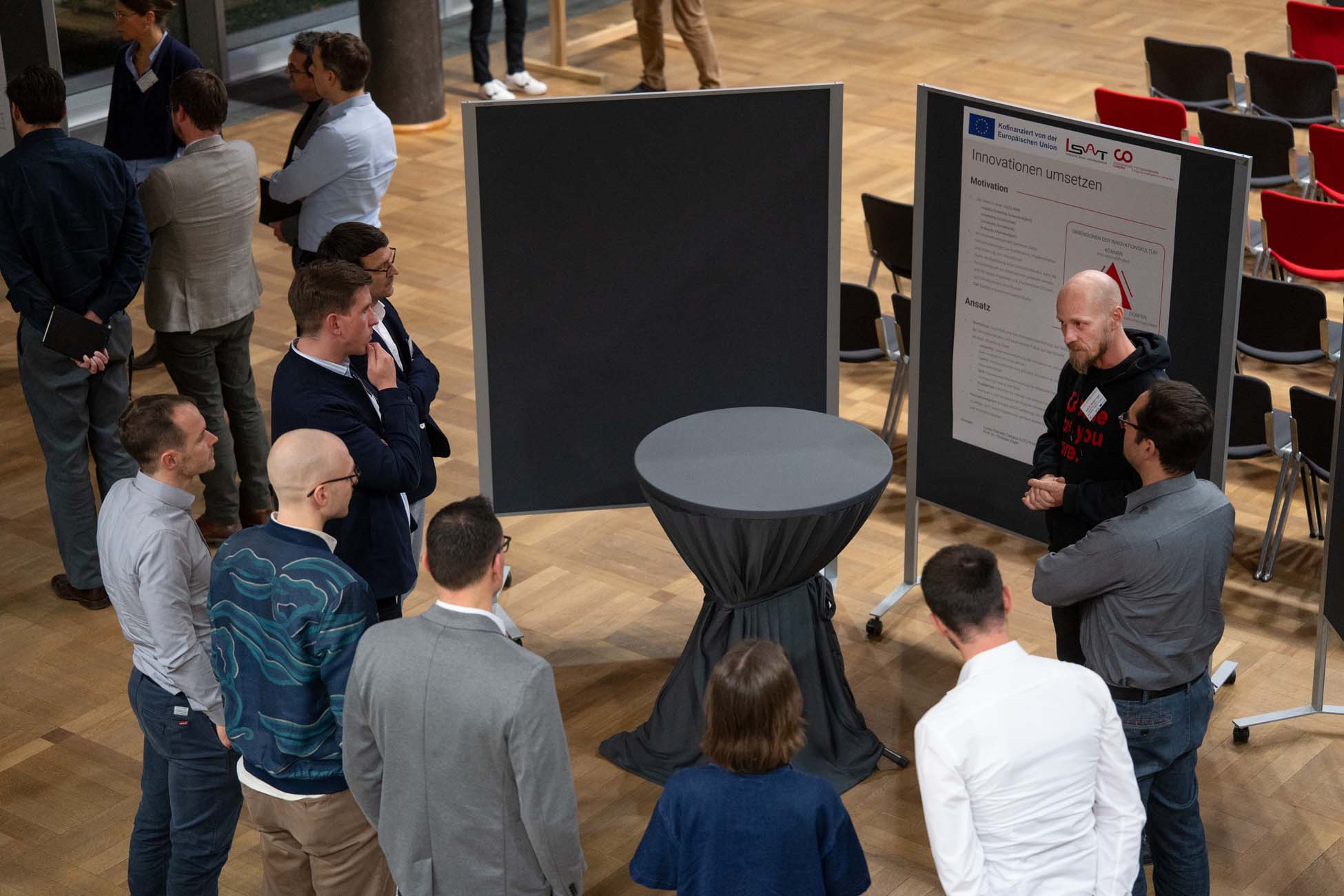 Eine Gruppe von Personen steht bei einer Veranstaltung in einem Gebäude um einen Stehtisch herum und diskutiert, wobei im Hintergrund Präsentationstafeln und Stühle zu sehen sind - vielleicht wird ein automatisch gespeicherter Entwurf besprochen. at Hochschule Coburg