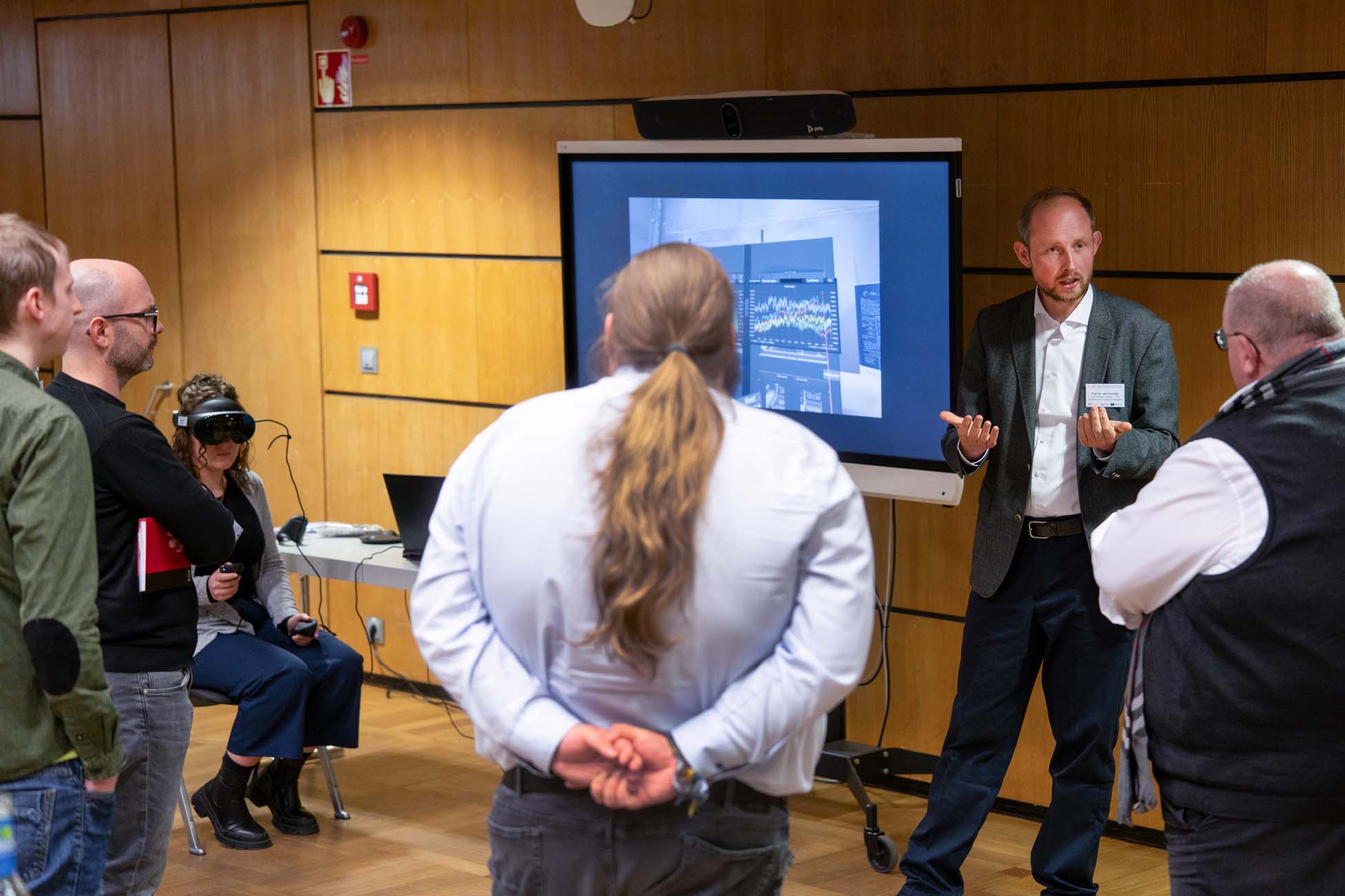 Ein Mann im Anzug präsentiert einer kleinen Gruppe in einem holzgetäfelten Raum Daten auf einem großen Bildschirm; in der Nähe konzentriert sich eine Person, die ein VR-Headset und Kopfhörer trägt, auf einen automatisch gespeicherten Entwurf auf ihrem Laptop. at Hochschule Coburg