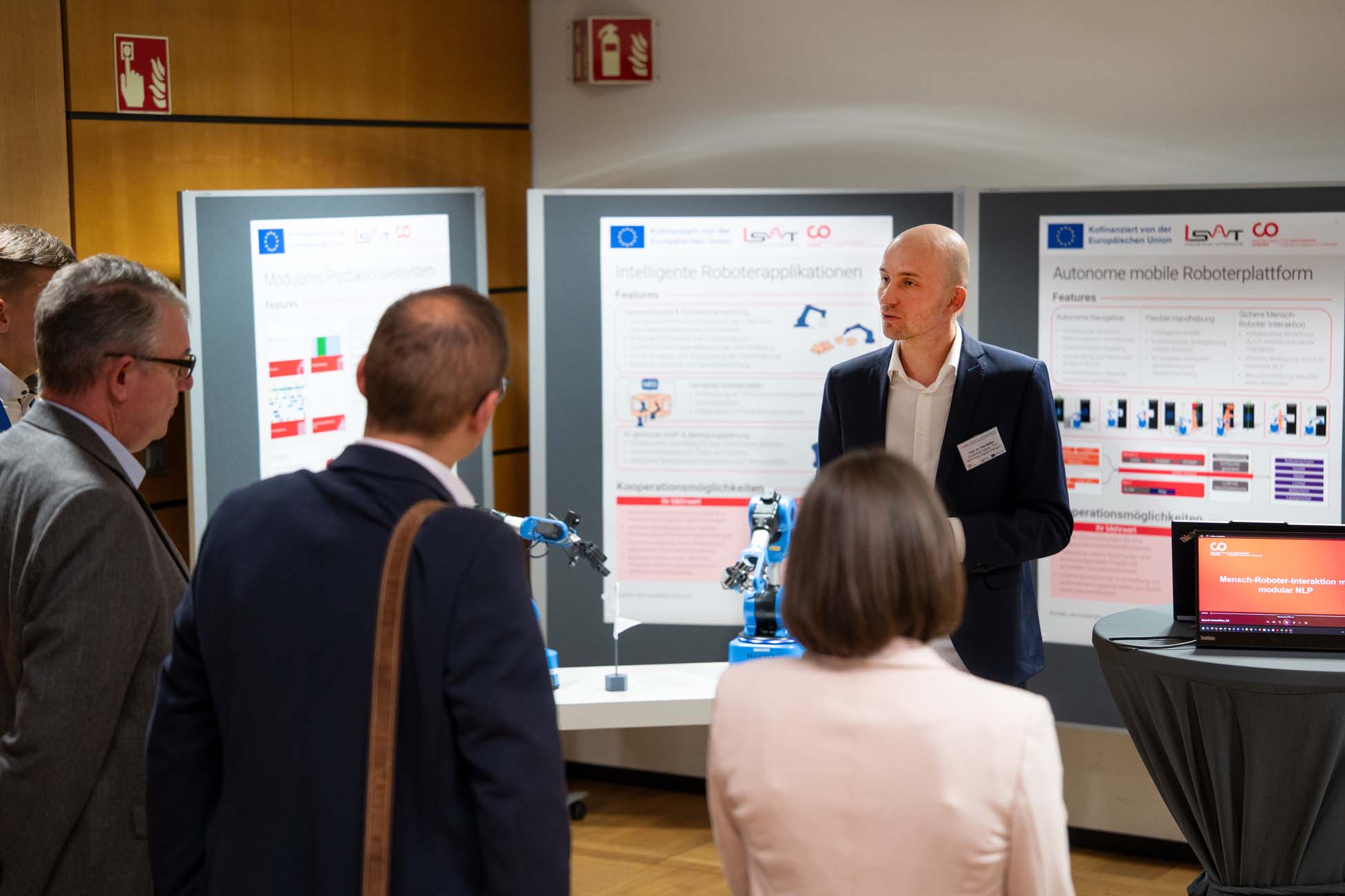 Ein Mann im Anzug präsentiert einer kleinen Gruppe in einem Ausstellungsraum Robotertechnologie. Hinter ihm sind Informationsplakate und ein Roboterarm zu sehen, der die neuesten Innovationen des Automatisch gespeicherten Entwurfs vorstellt. at Hochschule Coburg