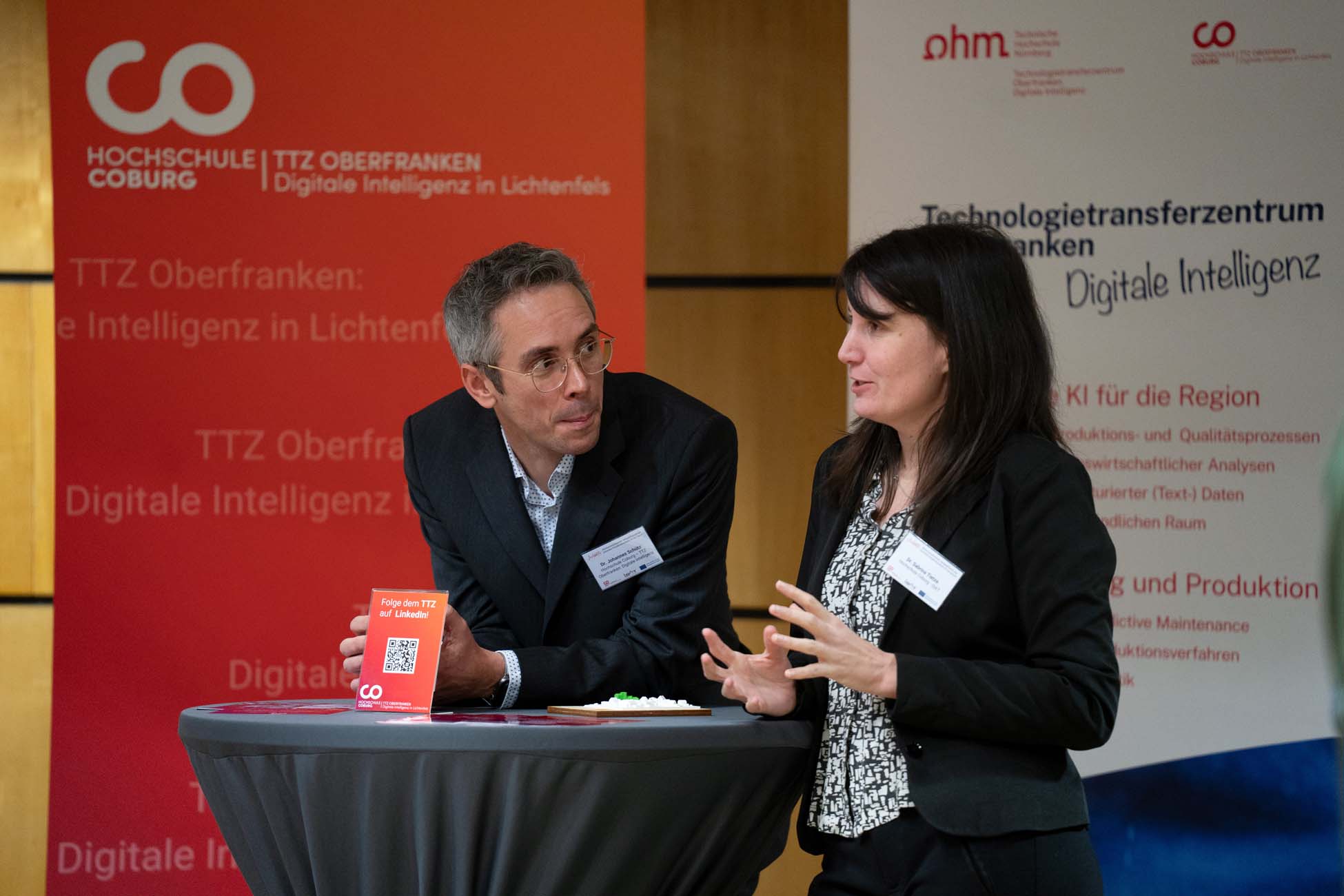 Zwei Fachleute stehen an einem runden Tisch und unterhalten sich auf einer Konferenz. Hinter ihnen hängen Transparente mit deutschem Text über digitale Intelligenz und Technologietransfer an der Hochschule Coburg: "Automatisch gespeicherter Entwurf. at Hochschule Coburg