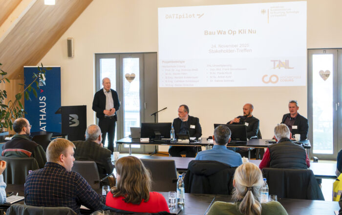 Ein Mann steht und spricht in einem hellen Konferenzraum zu einem Publikum über Landschaftsschutz, während drei andere Männer an einem Tisch sitzen, der dem Publikum zugewandt ist. Hinter ihnen ist eine große Präsentationsfolie zu sehen. Die Anwesenden sitzen und hören aufmerksam zu. at Hochschule Coburg