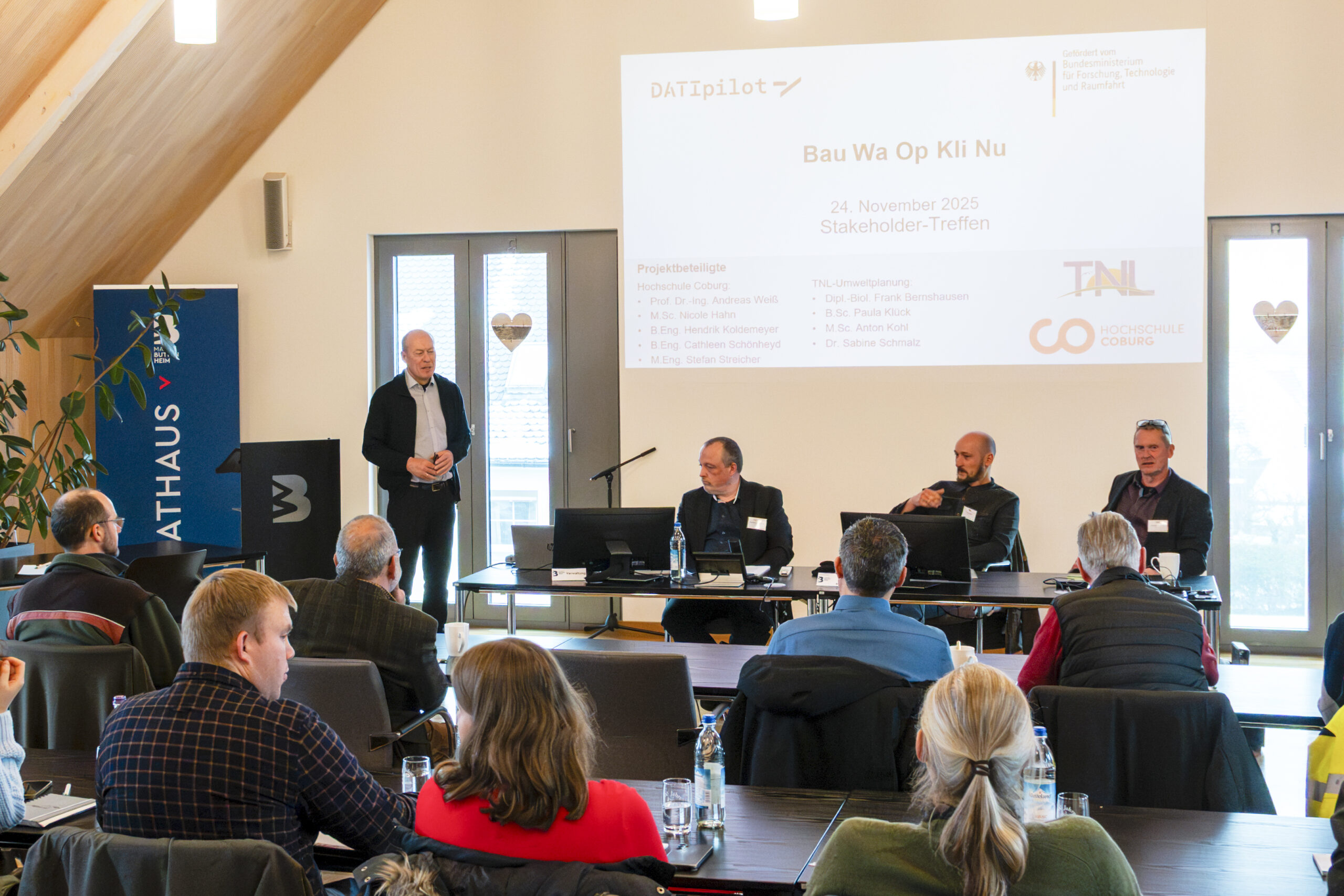 Ein Mann steht und spricht in einem hellen Konferenzraum zu einem Publikum über Landschaftsschutz, während drei andere Männer an einem Tisch sitzen, der dem Publikum zugewandt ist. Hinter ihnen ist eine große Präsentationsfolie zu sehen. Die Anwesenden sitzen und hören aufmerksam zu. at Hochschule Coburg