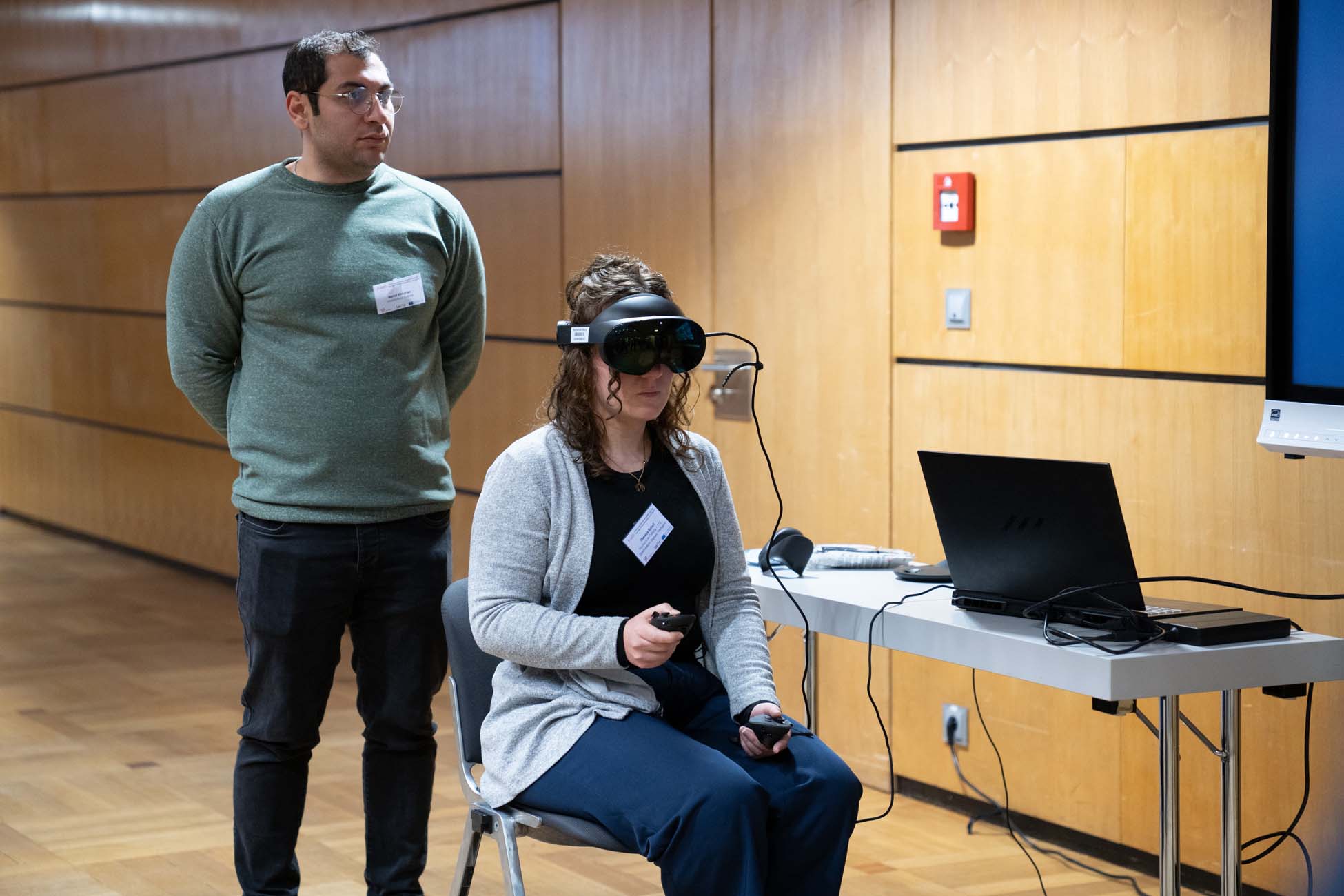 Eine Frau sitzt mit einem Virtual-Reality-Headset und hält einen Controller in der Hand, während ein Mann hinter ihr steht. In diesem Raum mit Holzwänden, neben einem Tisch mit einem Laptop und Kabeln, ist ein automatisch gespeicherter Entwurf auf dem Bildschirm geöffnet. at Hochschule Coburg