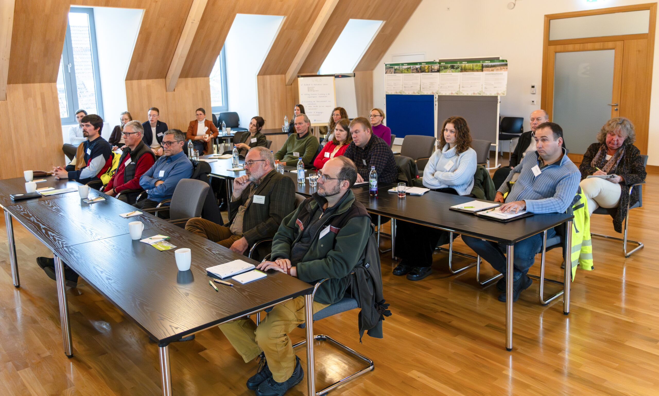 Eine Gruppe Erwachsener sitzt aufmerksam an Tischen in einem hellen, modernen Konferenzraum mit Holzböden und schrägen Decken und verfolgt einen Vortrag über Landschaftsschutz. Auf den Tischen liegen Notizbücher, Stifte und Tassen. at Hochschule Coburg