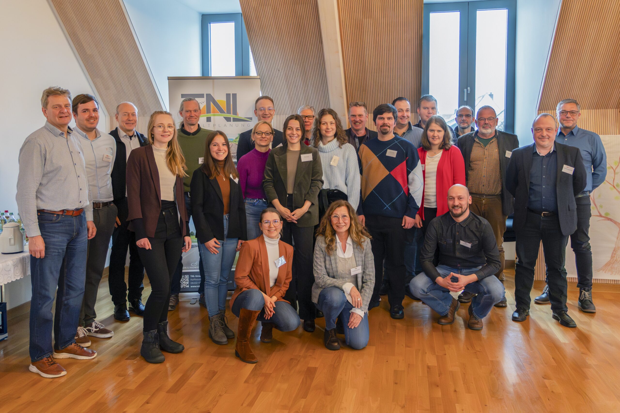Eine Gruppe von etwa 20 Personen posiert für ein Foto in einem hellen Raum mit Holzboden und schrägen Decken. In legerer Business-Kleidung stehen und knien sie vor einem Transparent zum Thema Landschaftsschutz und Fenster. at Hochschule Coburg