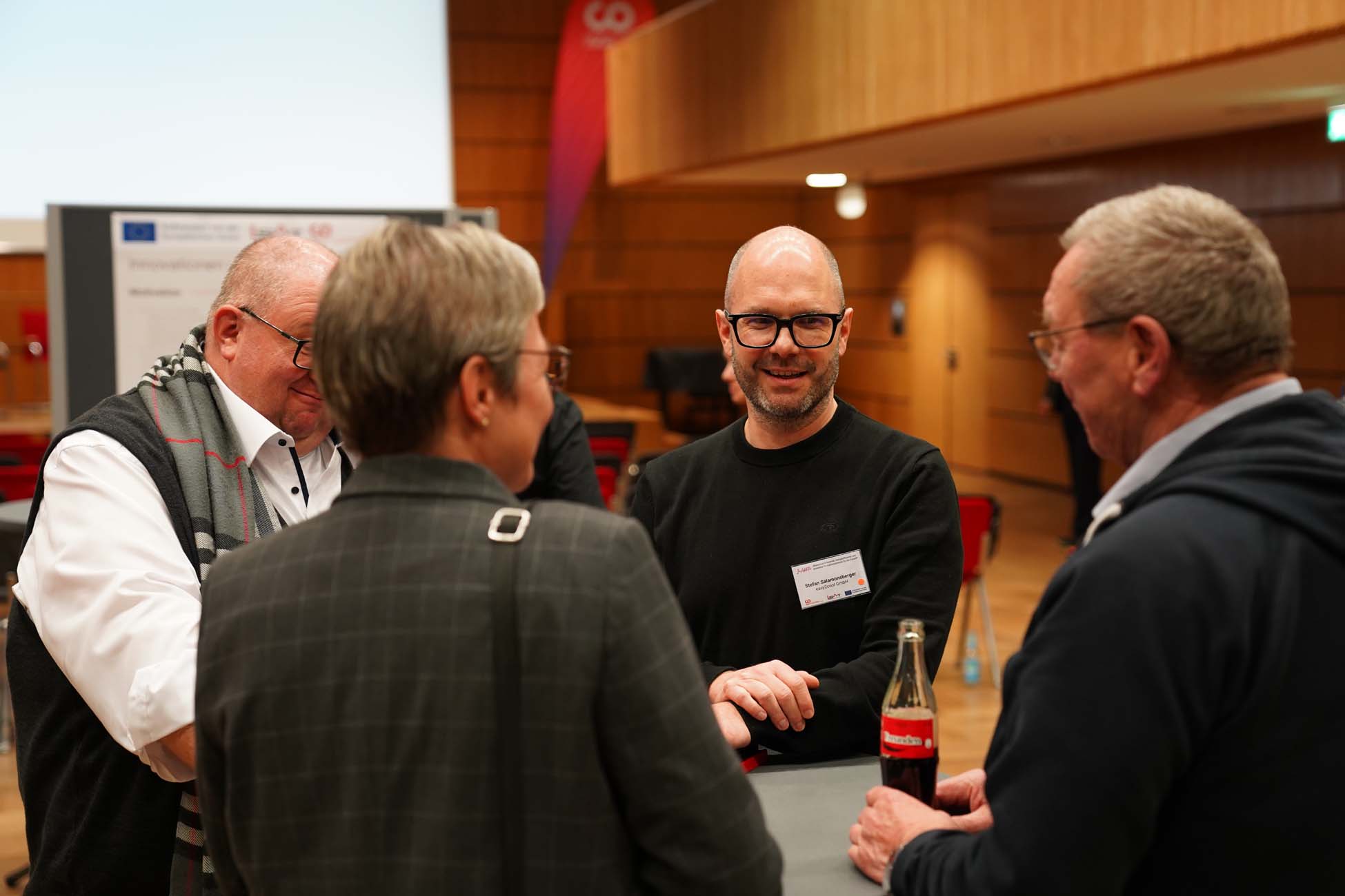 Vier Personen, drei Männer und eine Frau, stehen in einem Konferenzraum und unterhalten sich an einem Tisch. Ein Mann hält eine Coca-Cola-Flasche in der Hand, und alle scheinen in ein freundliches Gespräch über einen automatisch gespeicherten Entwurf vertieft zu sein. at Hochschule Coburg