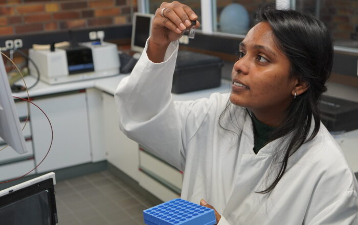 A scientist in a white lab coat examines a small vial closely while holding a blue test tube rack in a laboratory setting. Laboratory equipment and a brick wall are visible in the background. at Hochschule Coburg