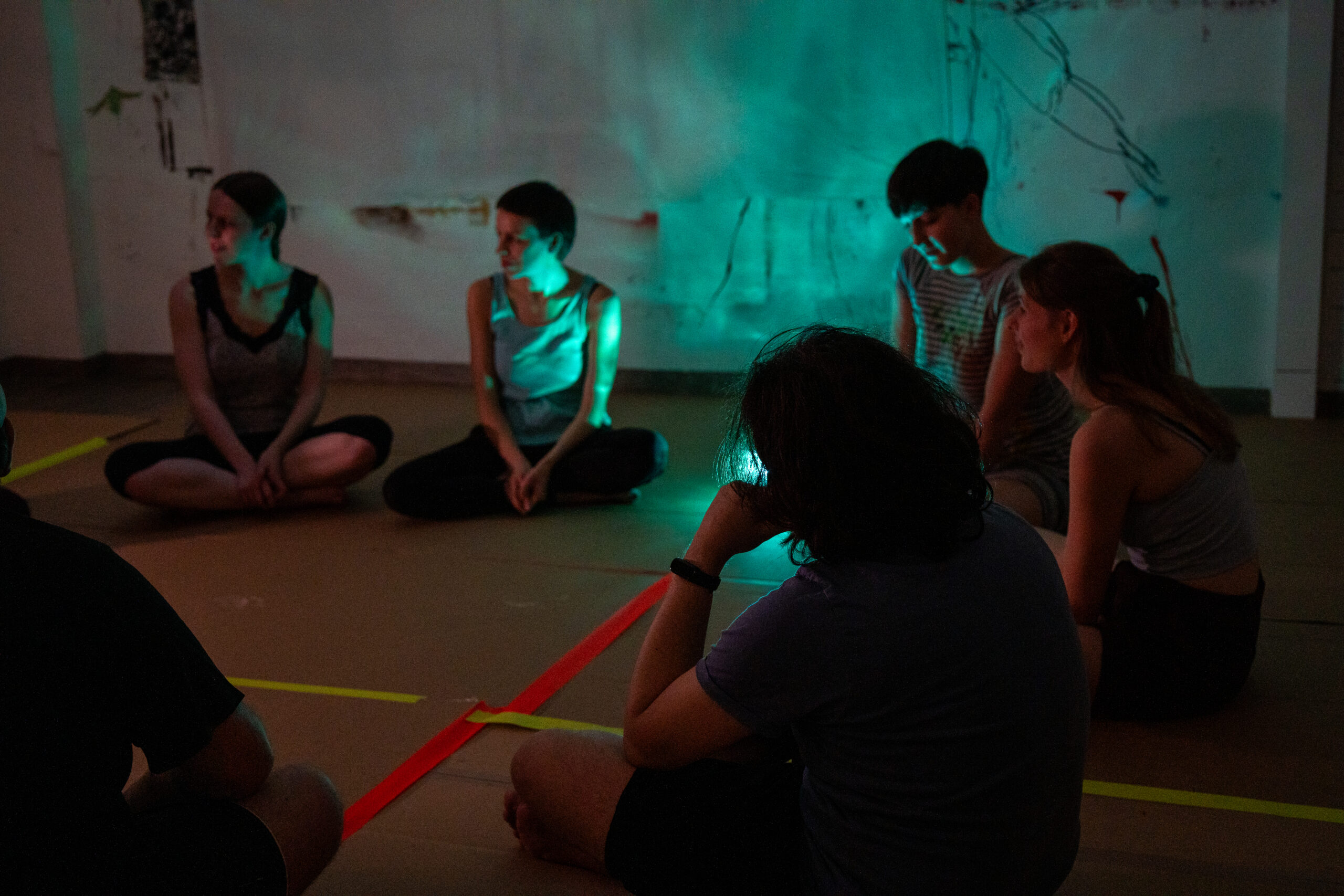 Fünf Personen sitzen im Schneidersitz auf einem mit gelbem und rotem Klebeband markierten Boden, der in einem mit Kunstwerken gefüllten Raum von schummrigem türkisfarbenem und grünem Licht beleuchtet wird. In dieser Umgebung, die an Kunst oder Angewandtes Theater erinnert, sitzen sie still und kontemplativ beisammen. at Hochschule Coburg