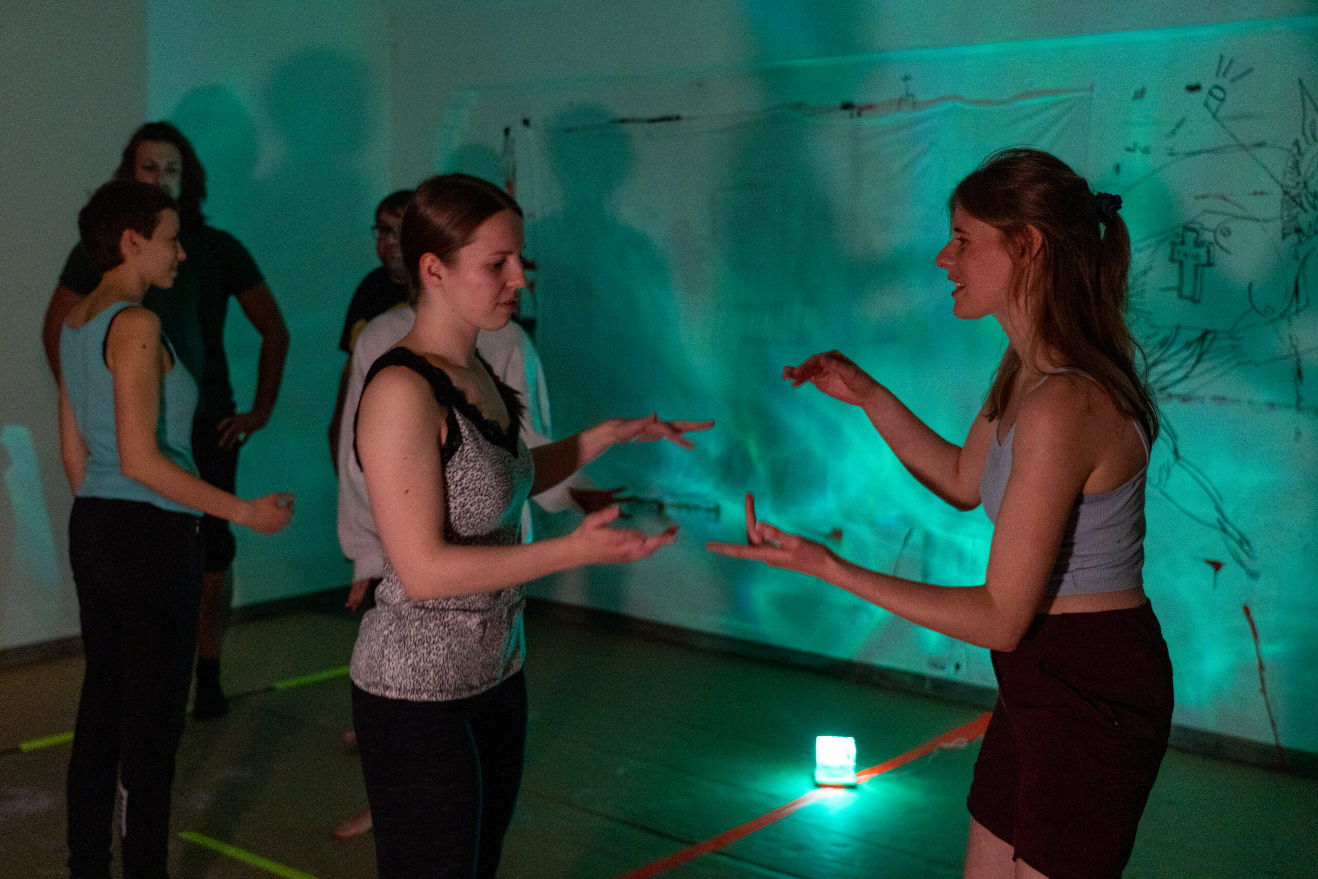 Mehrere Personen stehen in einem schwach beleuchteten Raum mit grünem Licht. Zwei Frauen im Vordergrund stehen sich gegenüber und gestikulieren wie bei einer angewandten Theaterübung oder einem Spiel, während andere zusehen. An der Wand hängen Zeichnungen, die der Szene einen Hauch von Kunst verleihen. at Hochschule Coburg