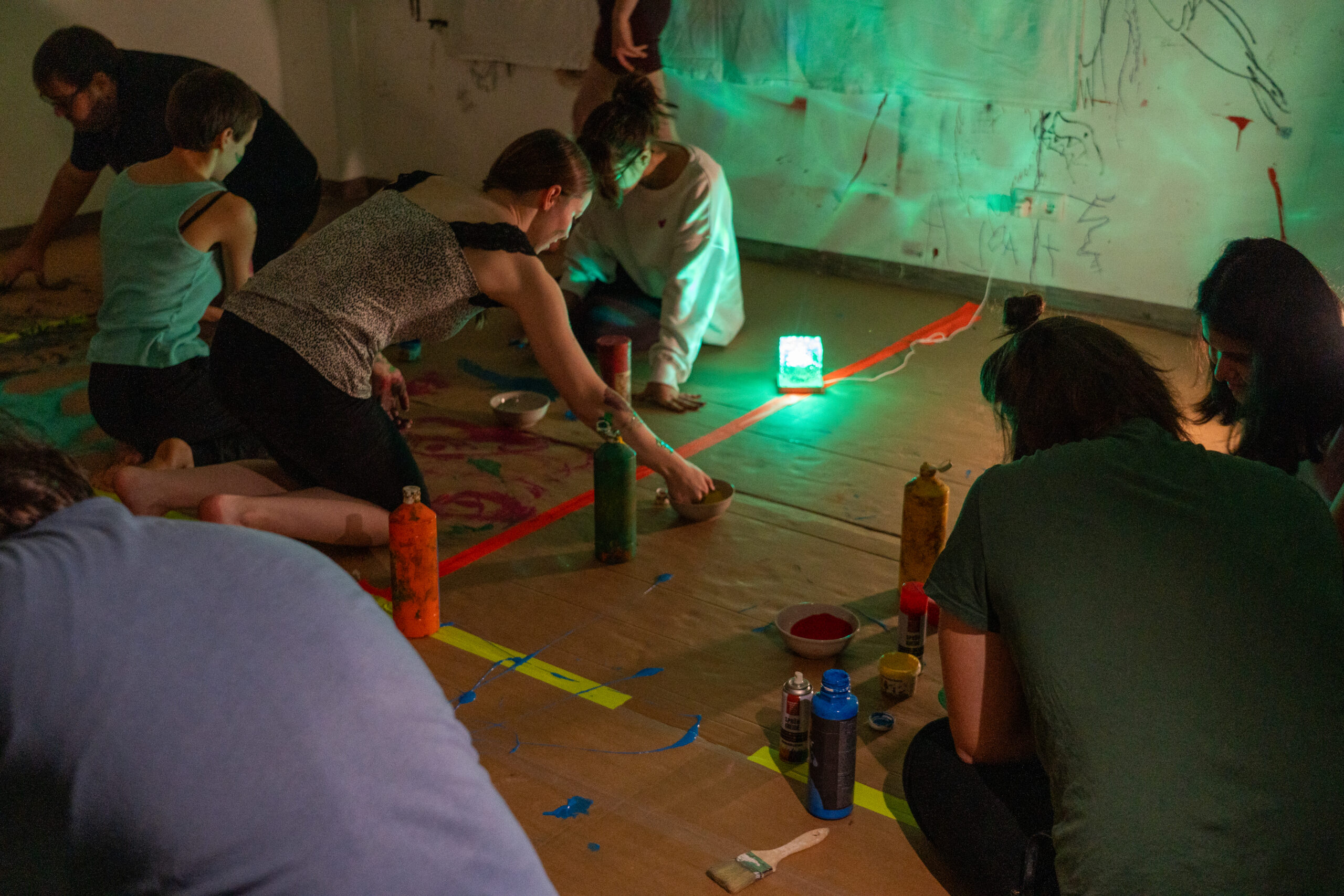 Mehrere Personen knien auf einem mit braunem Papier bedeckten Boden, umgeben von Farbflaschen und Pinseln, und schaffen unter grünem Umgebungslicht in einem schwach beleuchteten, mit Wandzeichnungen geschmückten Raum gemeinschaftliche Kunst. at Hochschule Coburg