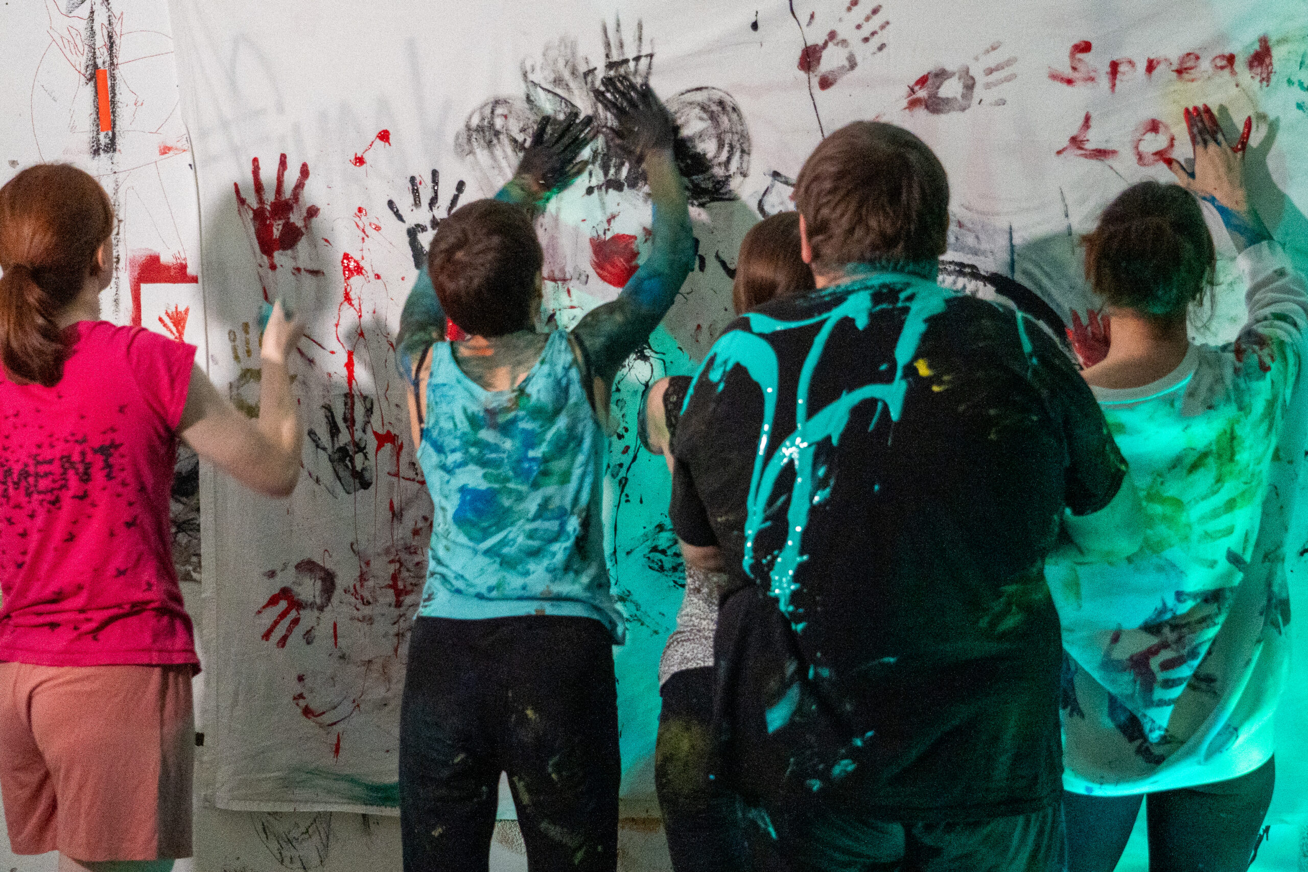 Fünf Personen mit farbverschmierten Kleidern drücken ihre Hände auf eine mit bunten Handabdrücken und Graffiti bedeckte Wand und hinterlassen weitere Spuren. Die von der Kunst inspirierte Szene ist kreativ und lebendig und wird von grünlichem Licht erhellt. at Hochschule Coburg