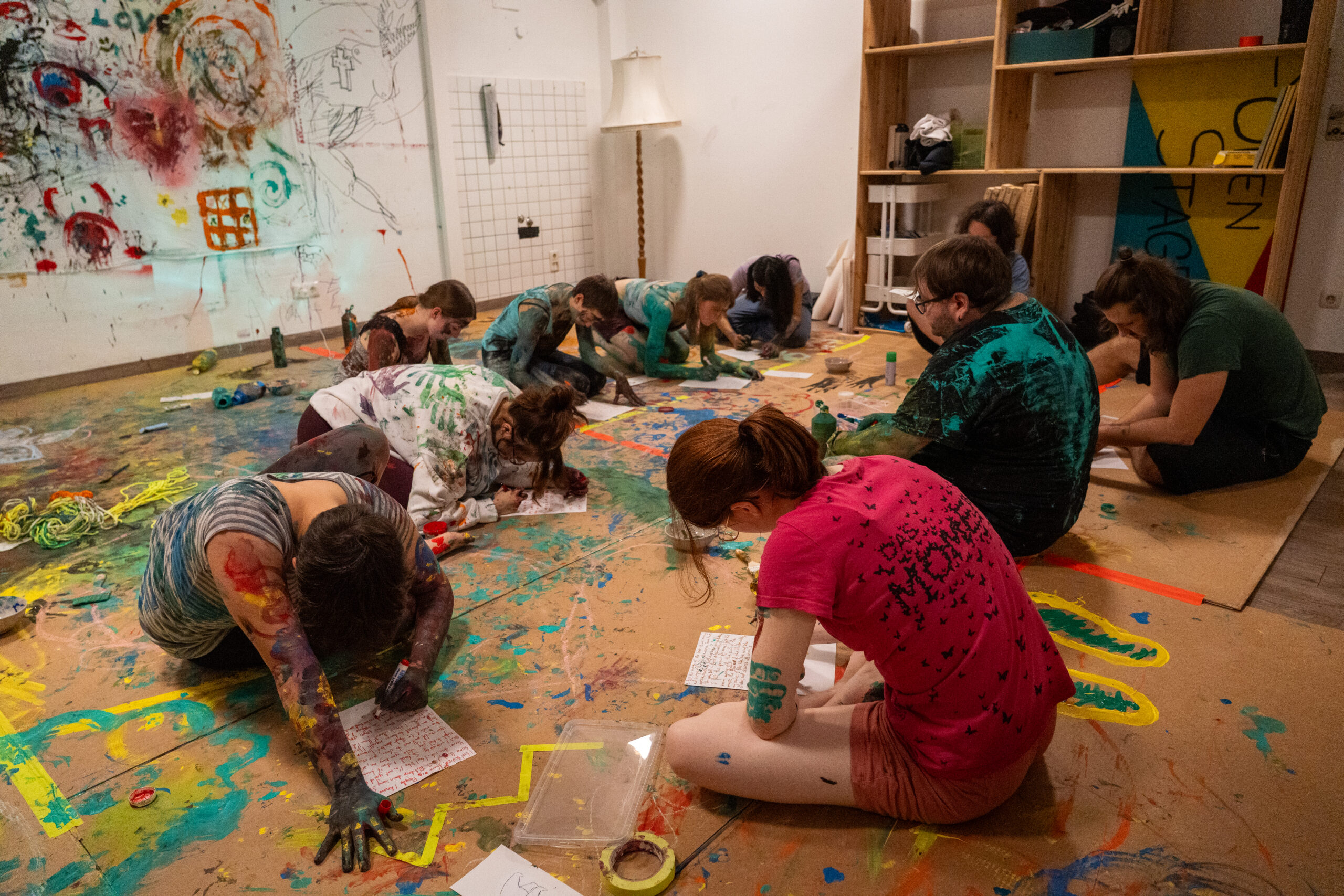 Eine Gruppe von Menschen, bedeckt mit bunter Farbe, sitzt und kniet auf einem unordentlichen Boden und arbeitet konzentriert an einzelnen Kunstprojekten. Der Raum ist mit Kunstzubehör übersät und bunte Farbspritzer bedecken den Boden und die Wände. at Hochschule Coburg
