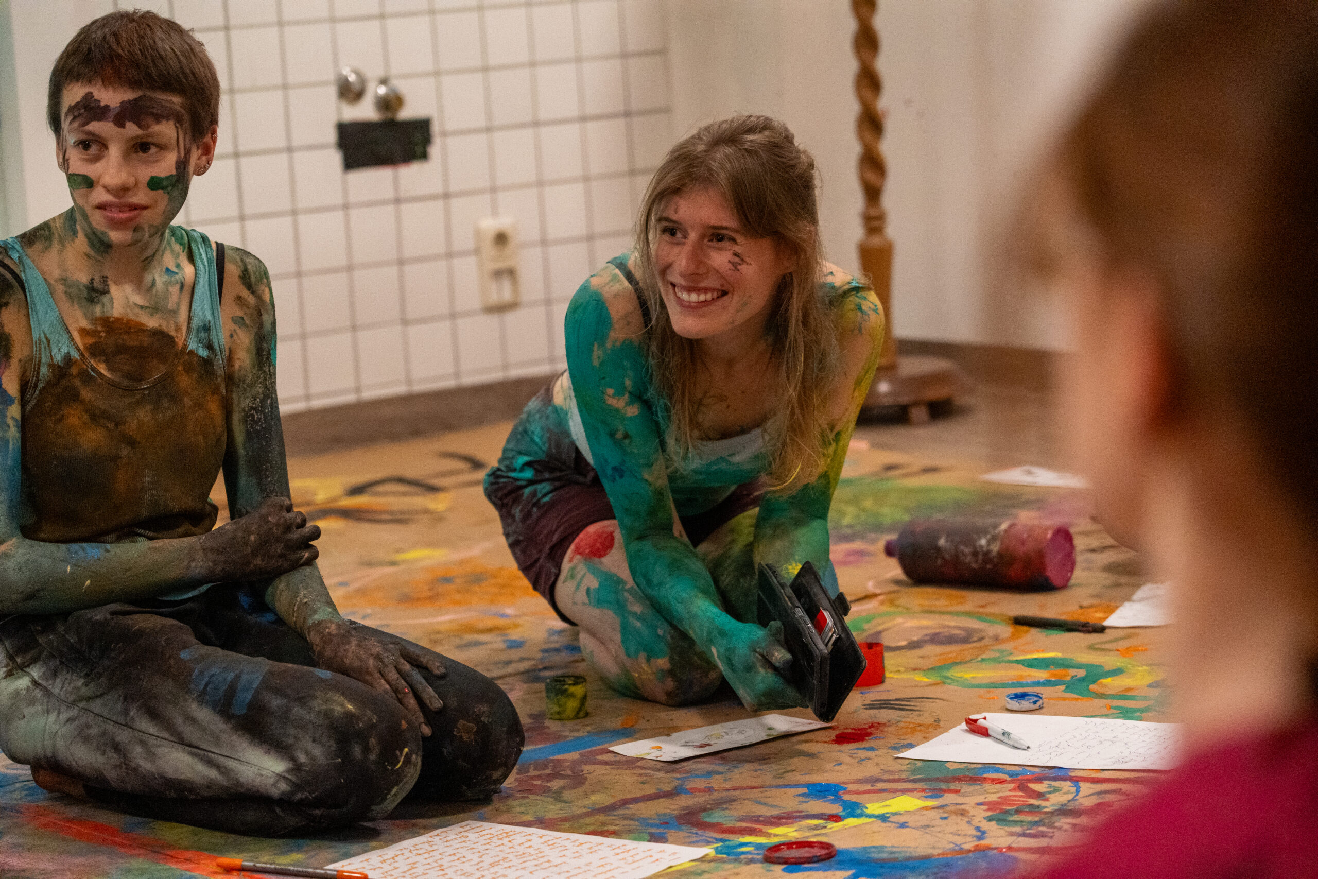Zwei mit bunter Farbe bedeckte Personen sitzen und knien auf einem mit Farbe bespritzten Boden und lächeln, während sie sich mit Papier und Kunstzubehör kreativ beschäftigen. Im Hintergrund sind eine geflieste Wand und verschiedene Kunstmaterialien zu sehen. at Hochschule Coburg
